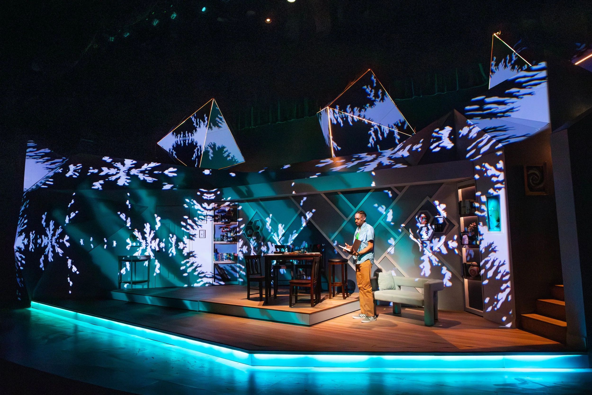 The image shows a stage with a person reading or working on a laptop, surrounded by colorful lighting and projected snowflake patterns.
