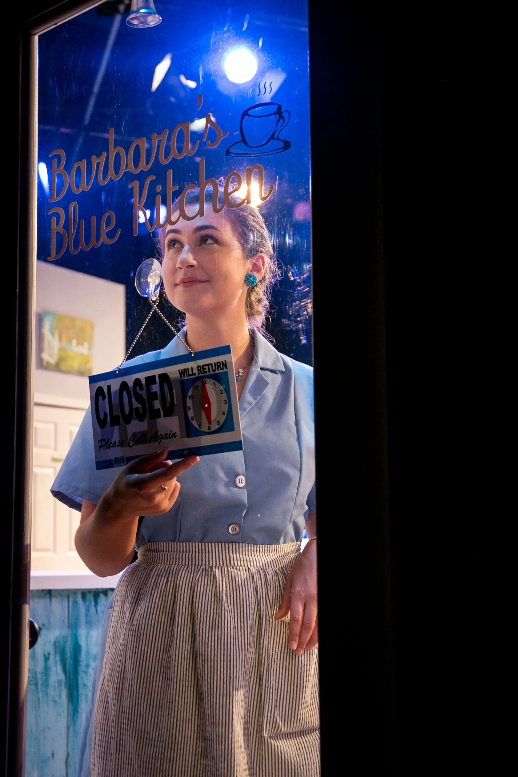 A woman dressed as a waitress or cook standing behind a glass door with a neon sign that reads 'Barbara's Blue Kitchen.' She is holding a sign that says 'CLOSED, WILL RETURN, Please Call Again' with a clock graphic showing 12 o'clock. The woman is lo