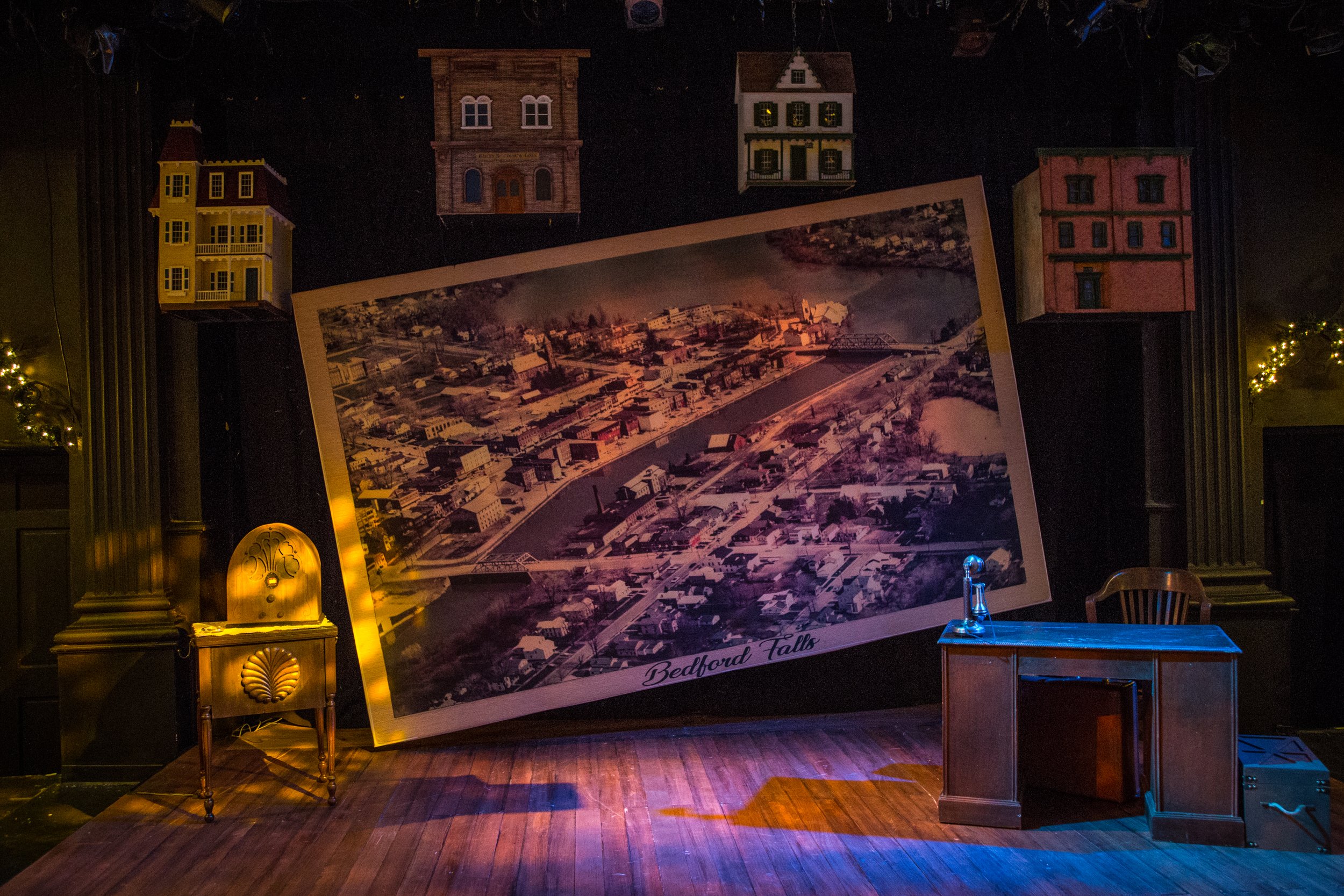 Stage setup with a large aerial photograph of Bedford Falls, leaning against a black backdrop, surrounded by miniature houses and a wooden desk with a glass lamp.