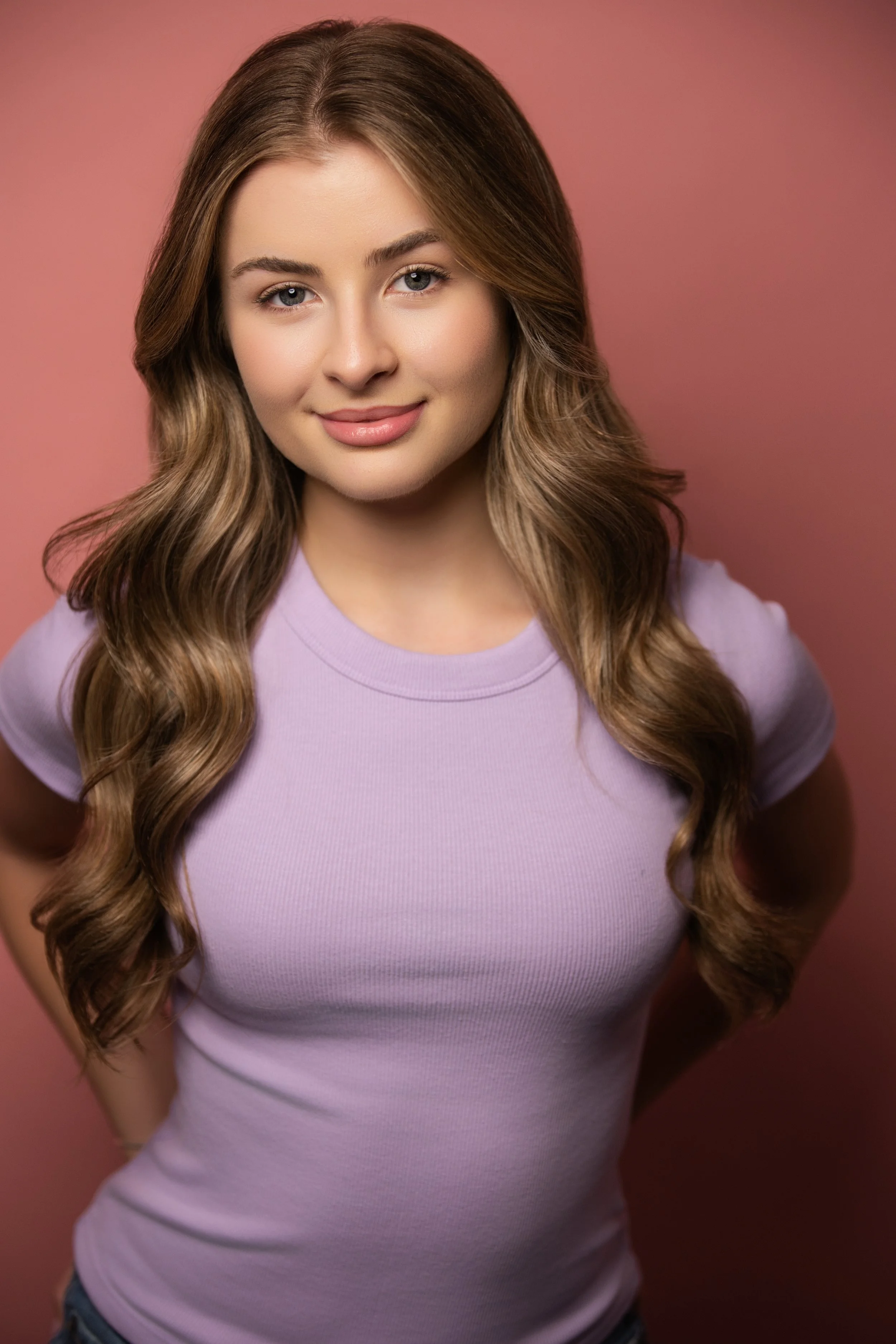 A young woman with long, wavy brown hair, wearing a light purple T-shirt, smiling slightly, standing against a pink background.