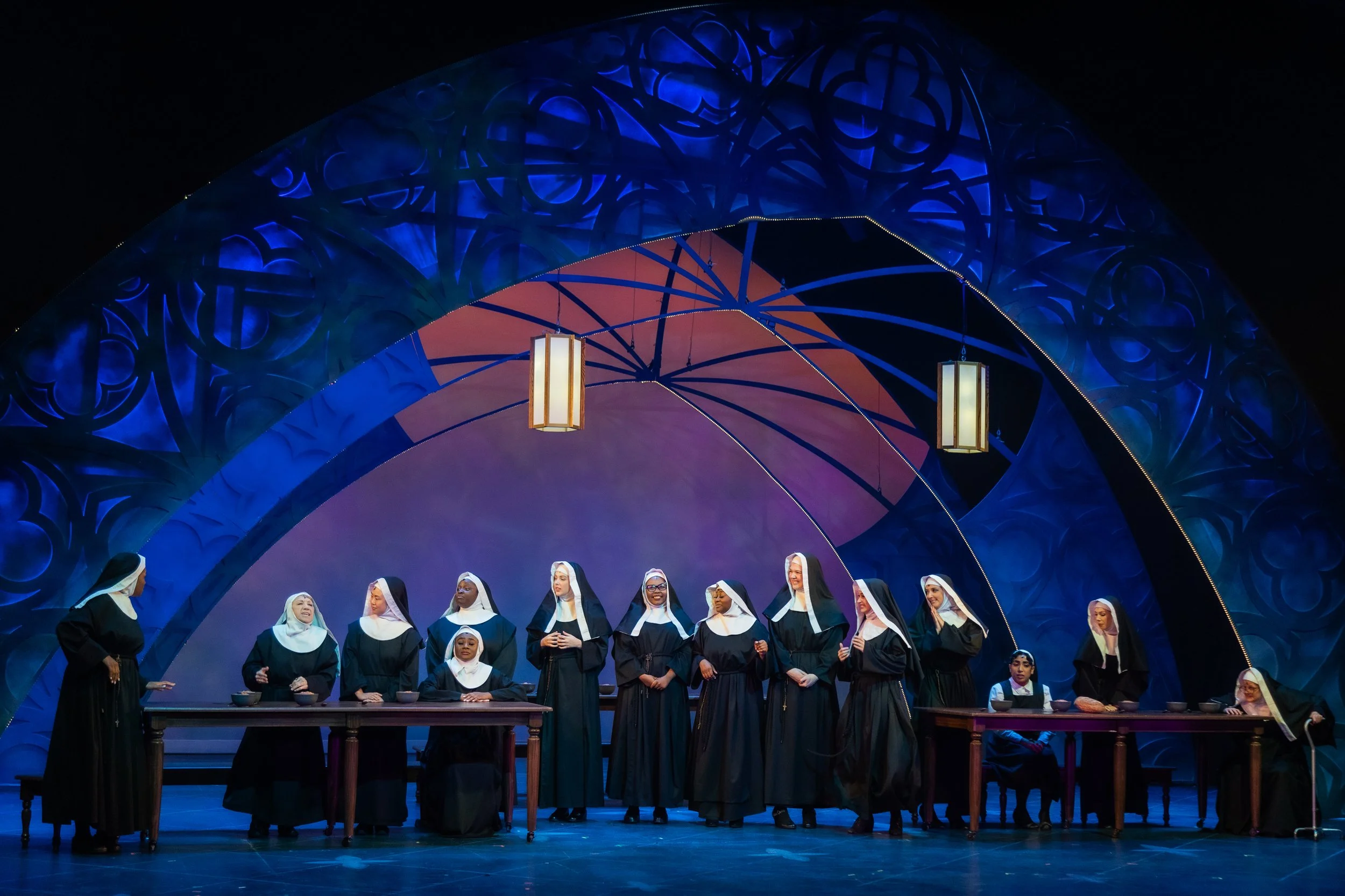 A theatrical stage depicting a group of nuns in black and white habits performing in a choir or musical ensemble, with a colorful and ornate backdrop and hanging lanterns above.