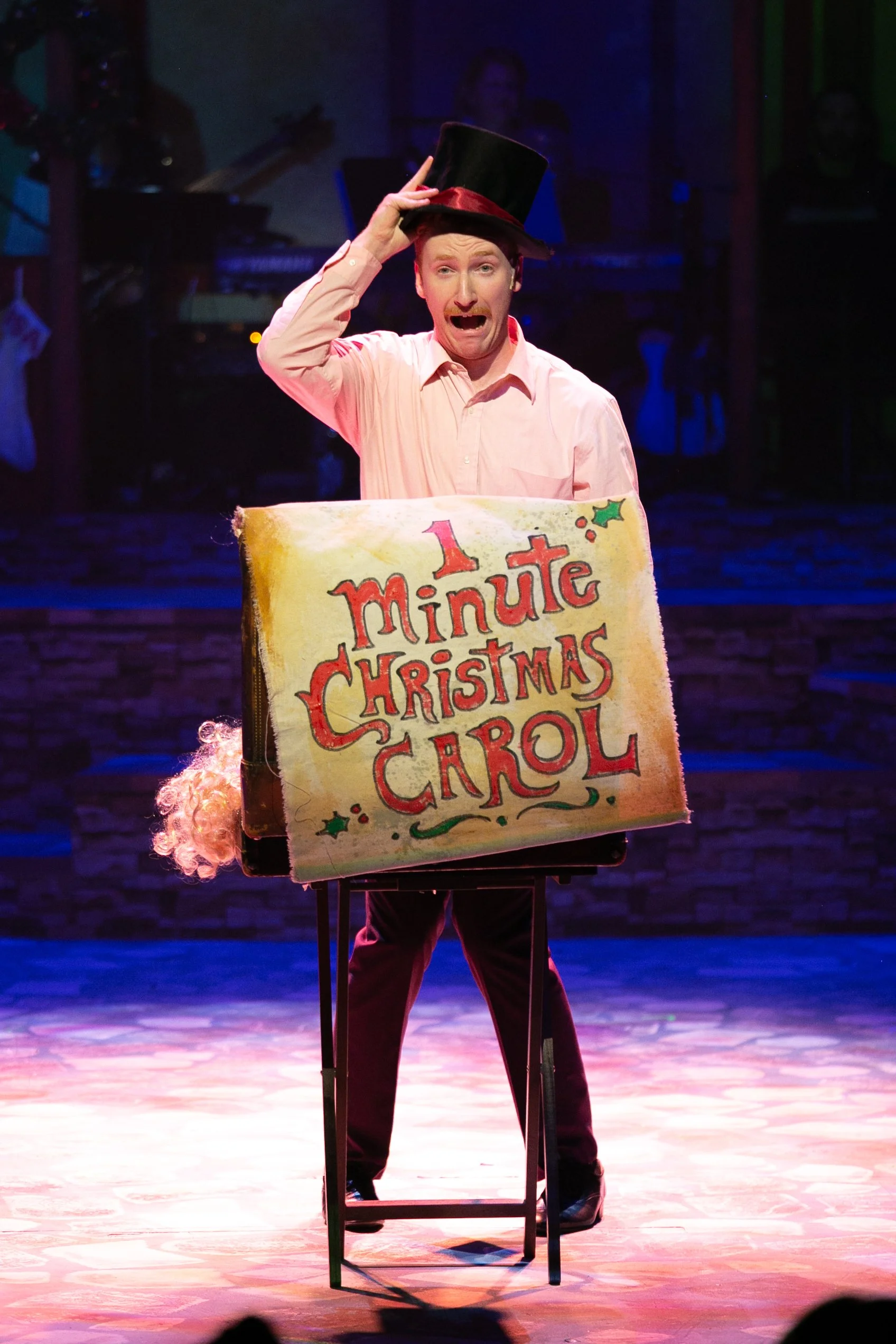 Performer holding a sign that reads '1 Minute Christmas Carol' on stage with a top hat, with a banjo and a microphone stand in the background.