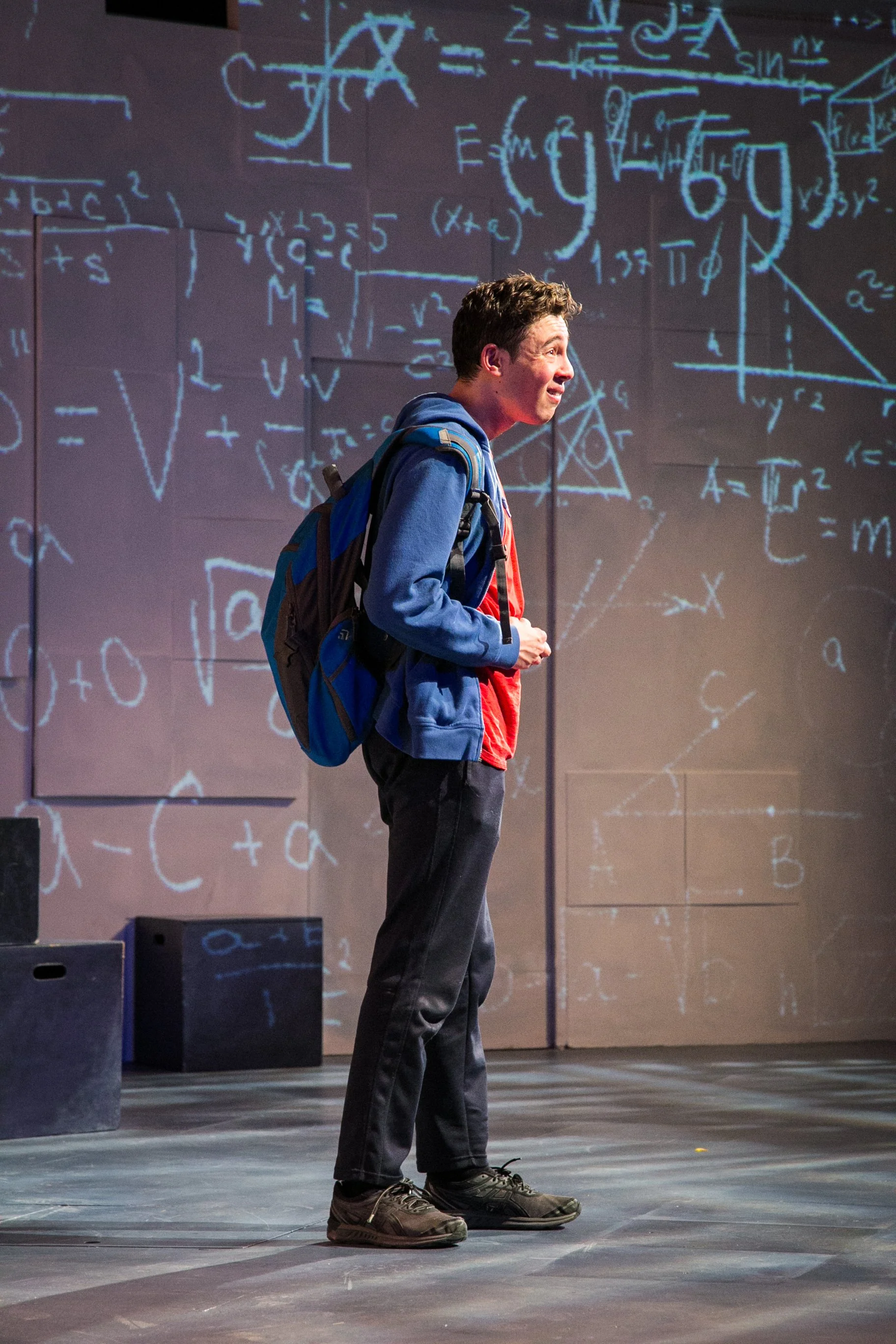 A young man with a backpack standing on a stage, speaking or singing, with a background of large chalkboard-like panels covered in scientific equations and diagrams.