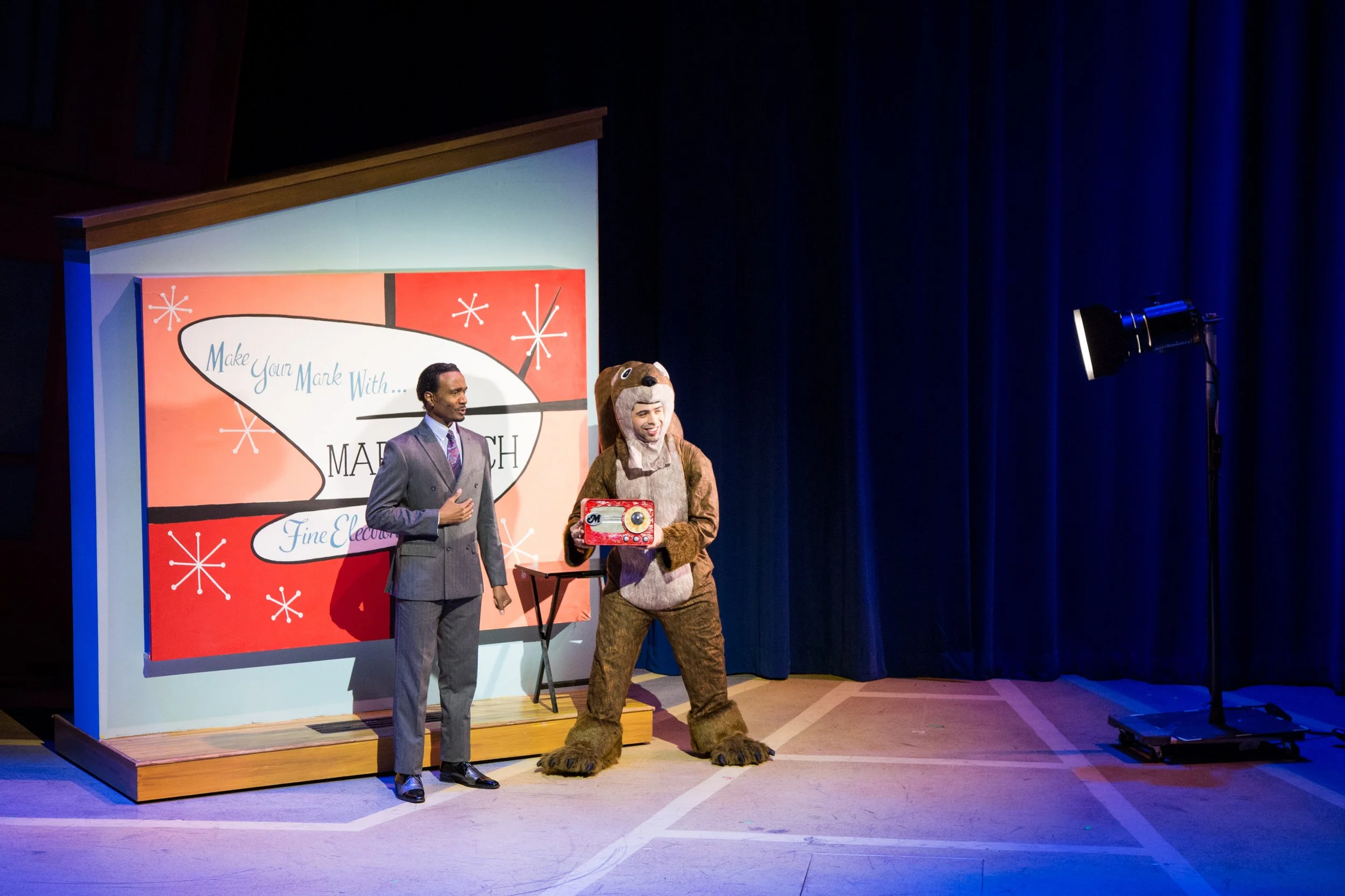 Two men on stage, one in a gray suit and the other in a bear costume, standing in front of a large red and blue backdrop with the text 'Make Your Mark With... Medium Fine Electronics', with stage lighting on the right.