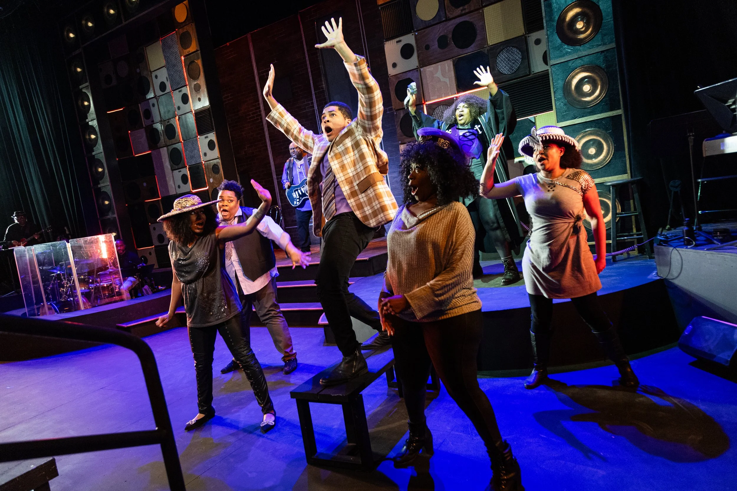 Actors performing on stage with various vocal and dance moves; colorful background with large speakers; musical instruments on side stage.
