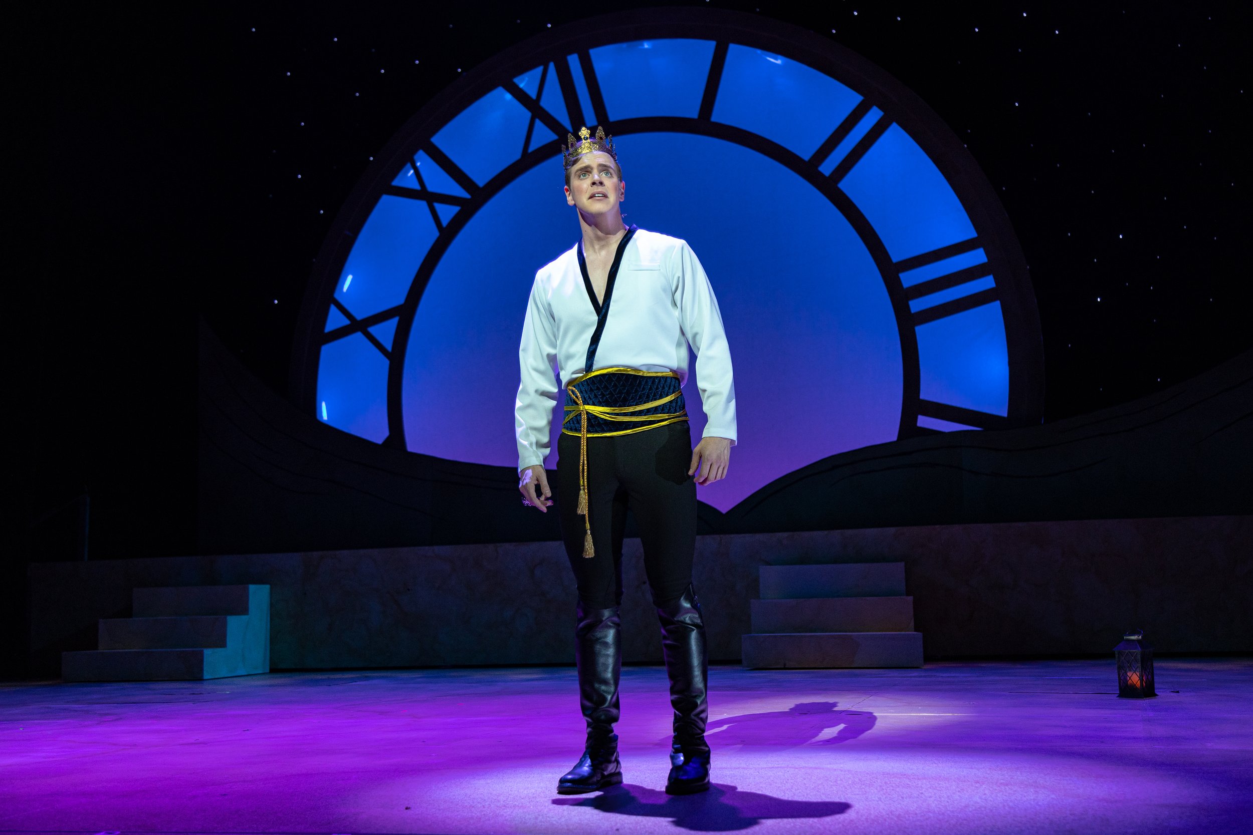 A young man wearing a white shirt, black pants, and tall black boots, with a blue and gold sash, stands on a stage in front of a large illuminated clock face, wearing a crown and looking upward, during a theatrical performance.