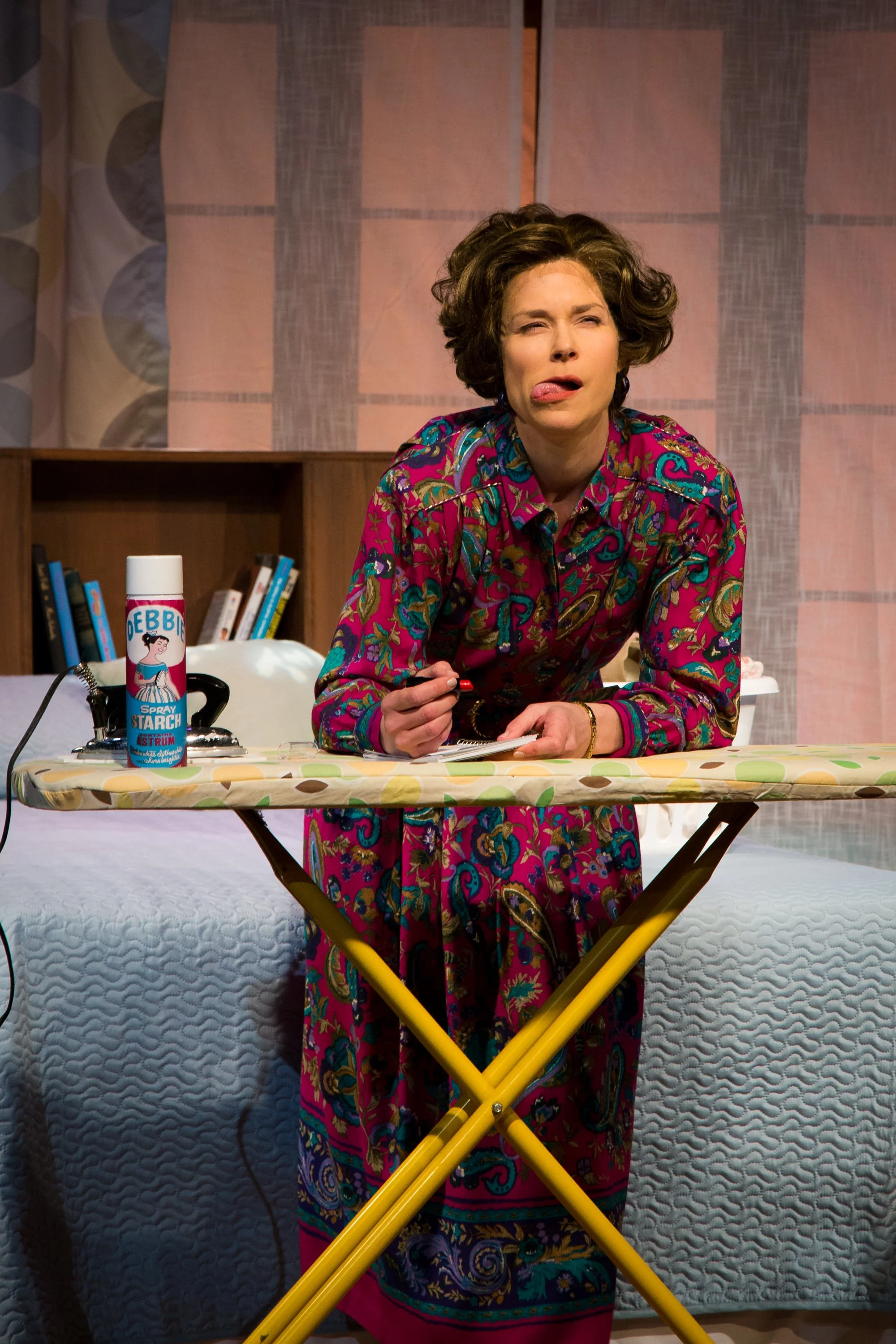 Woman with messy hair wearing a colorful patterned dress making a funny face with tongue out, leaning on an ironing board with a can of Debbie spray starch on it.