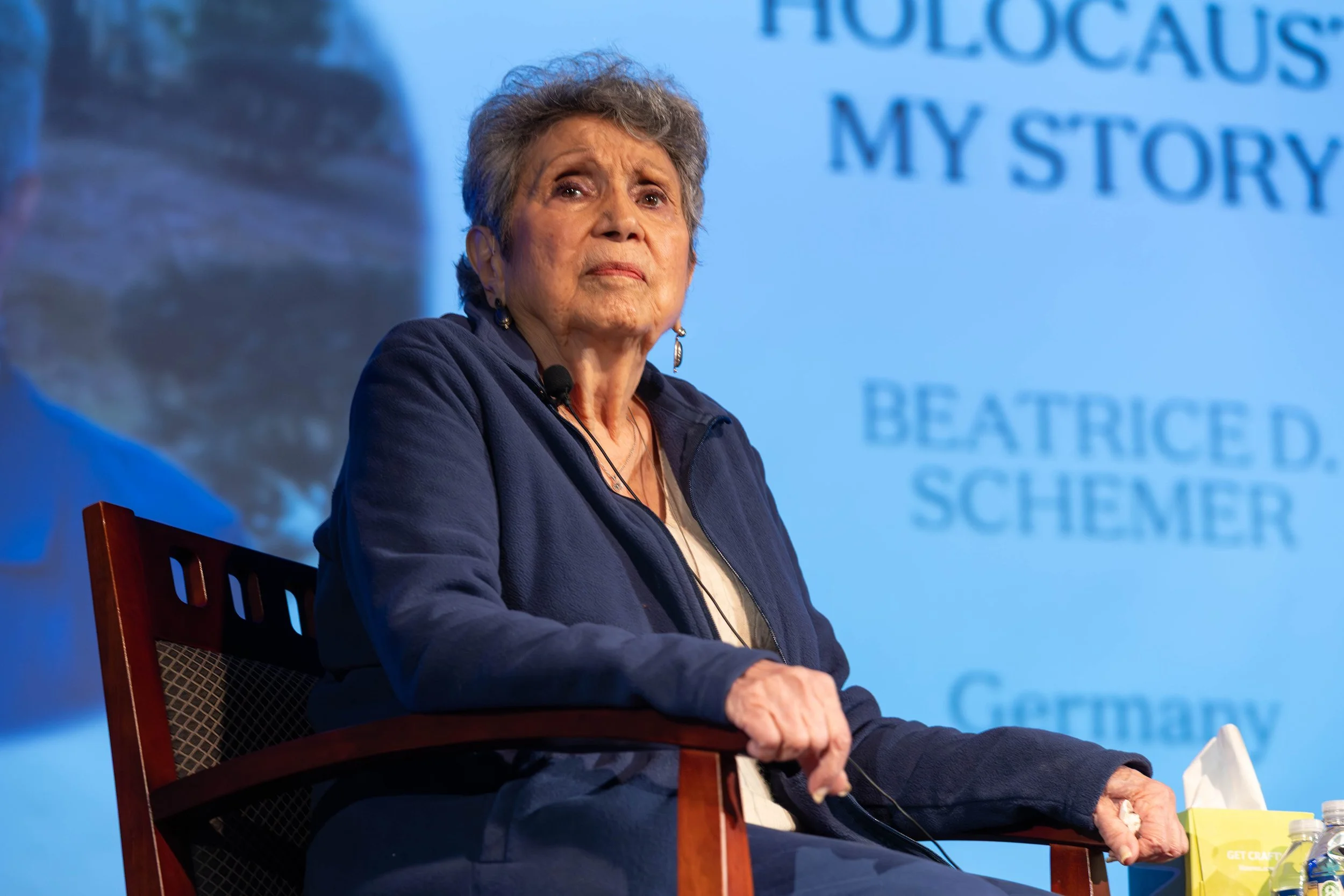 An elderly woman with gray hair sitting on a chair on a stage, wearing a navy blue jacket and earrings, with a projected background showing her name and storytelling about her Holocaust experience.