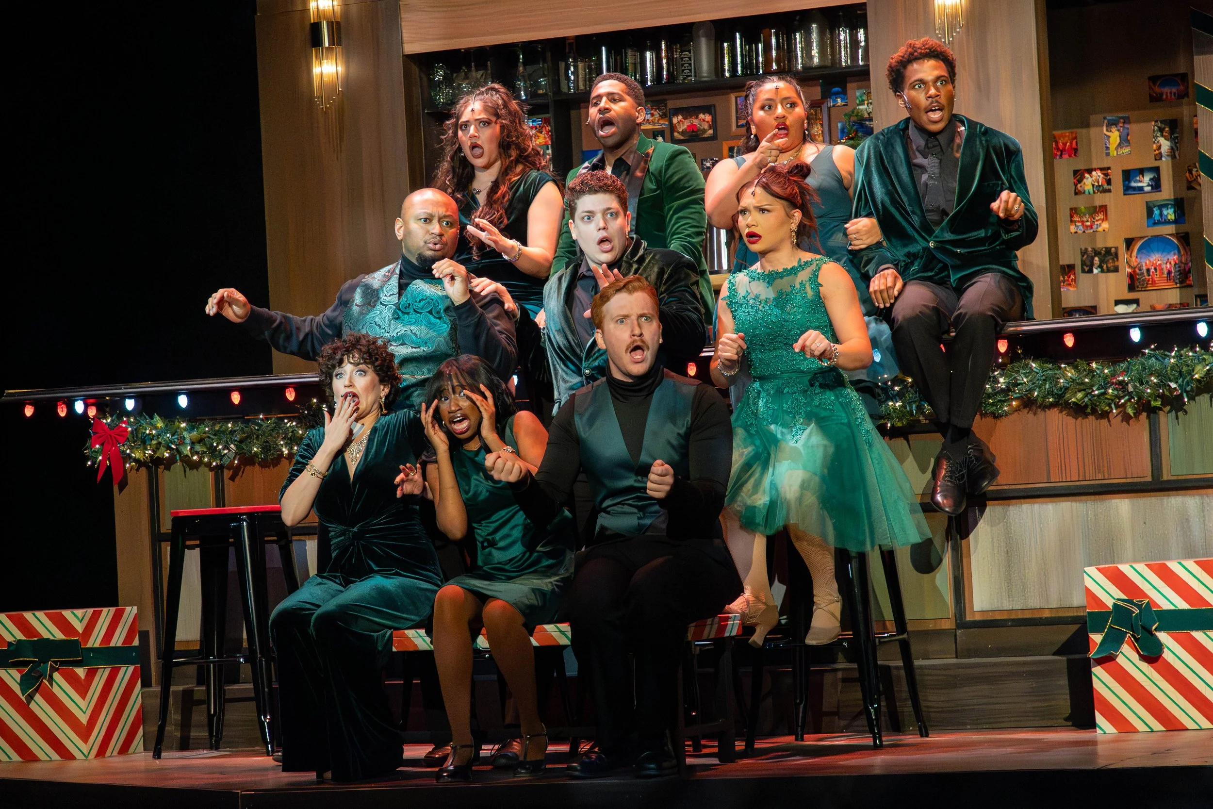 A group of people on stage with surprised and shocked expressions, sitting and standing on a set decorated with Christmas garland and presents, indicating a theatrical performance during the holiday season.
