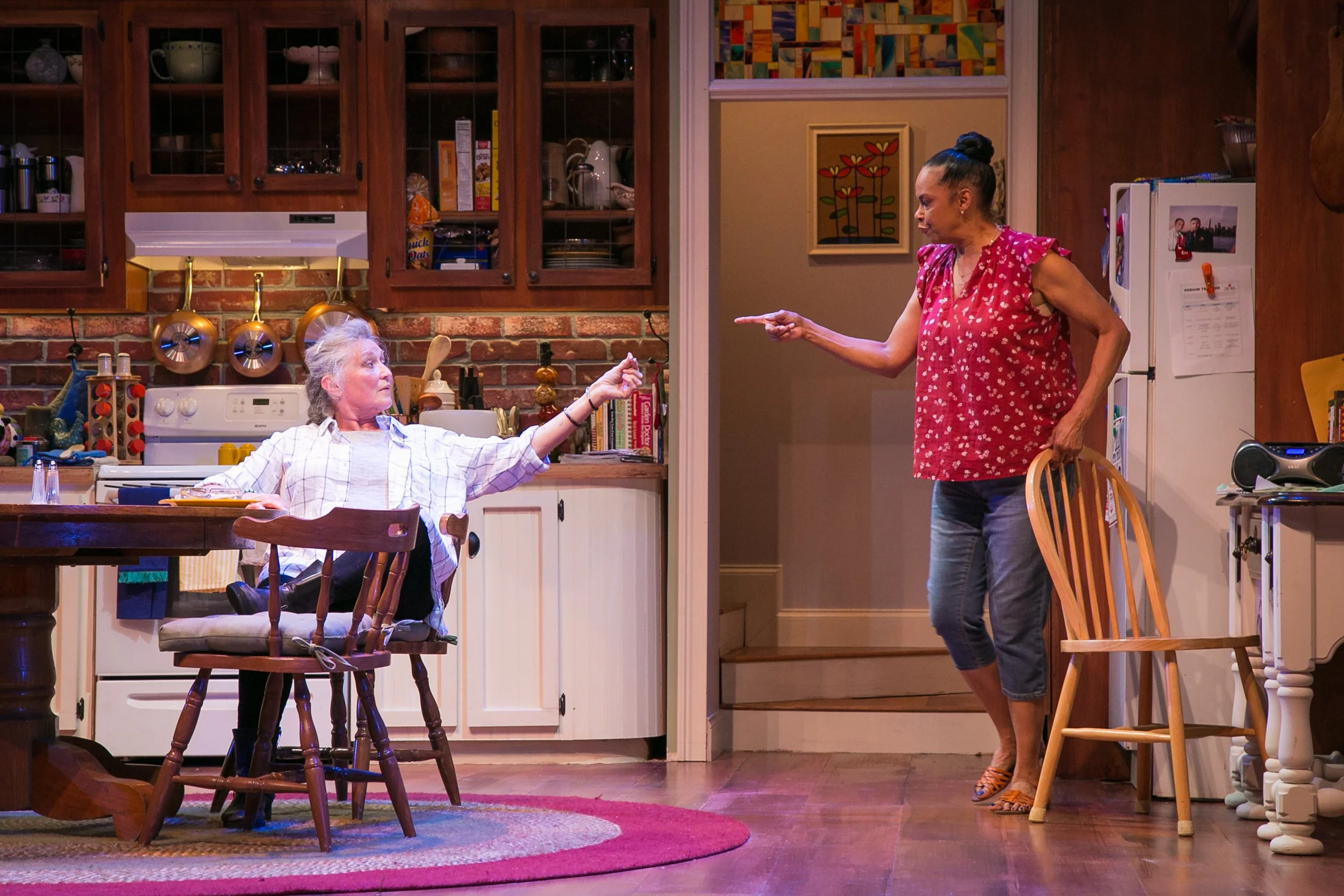 An older woman with gray hair sitting in a high chair in a kitchen, reaching out towards another woman with dark hair in a bun, wearing a red sleeveless top, who is standing nearby and pointing at her. The kitchen has wooden cabinets, a brick backspl