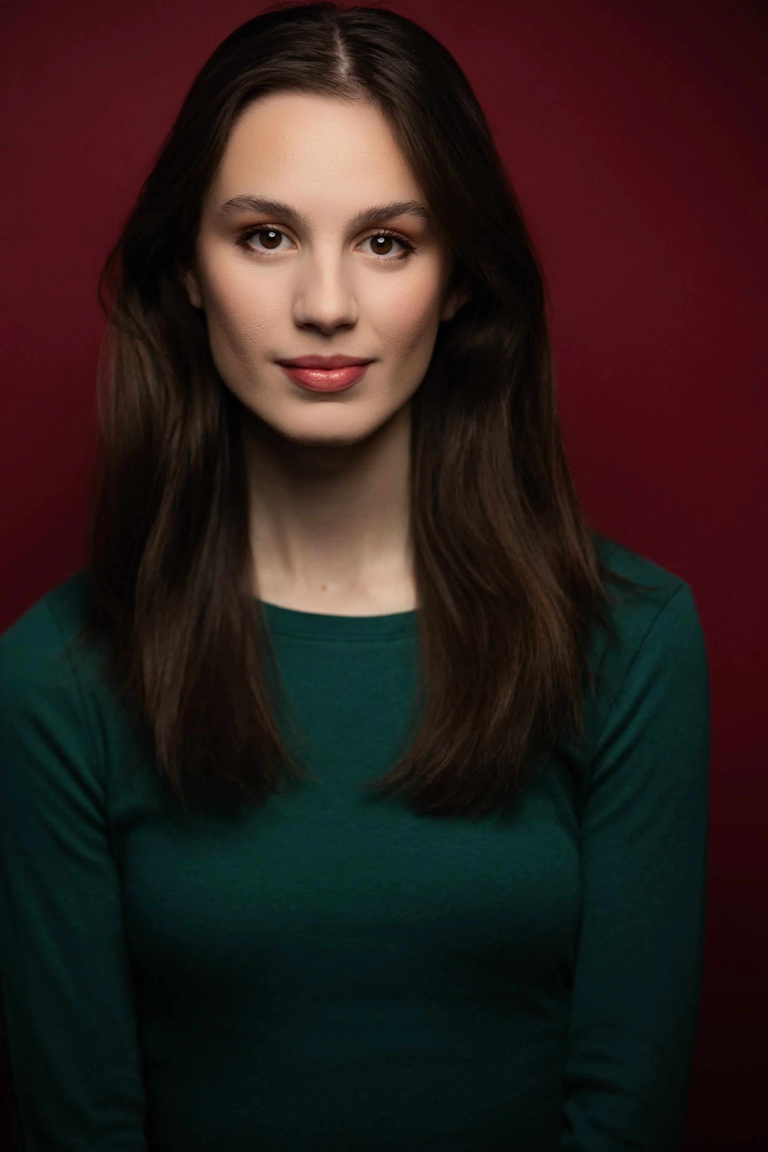 Portrait of a young woman with long brown hair, wearing a green shirt, smiling softly against a dark red background.