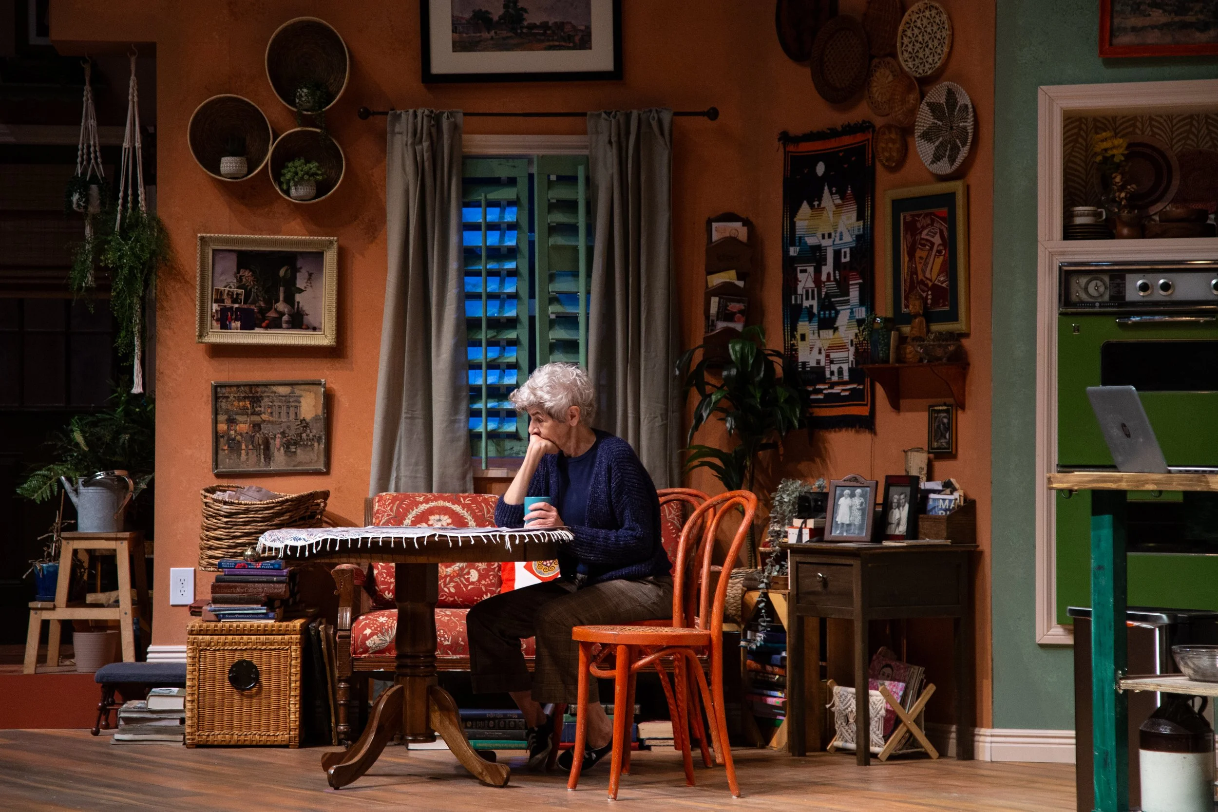 An elderly woman with white hair sitting at a dining table, holding a mug, looking pensive. The room is warmly lit with eclectic decor including framed art, hanging baskets, and a window with closed shutters. The space appears cozy and lived-in.