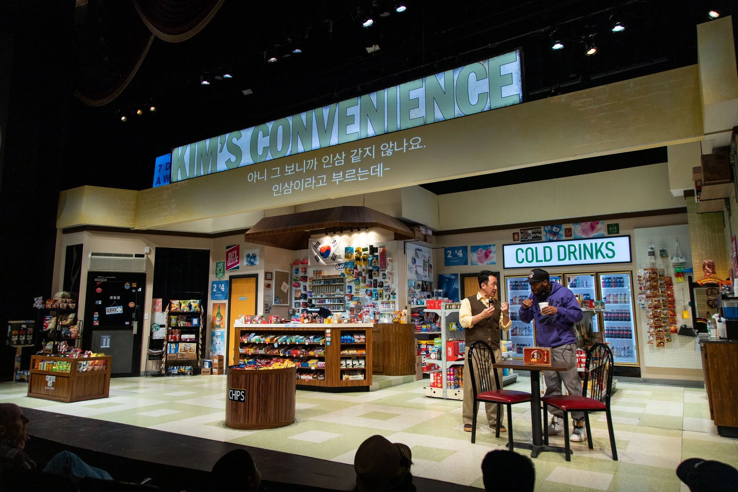 Stage set at a play depicting a store with a sign that says 'COLD DRINKS' and two actors standing and talking in front of the store.