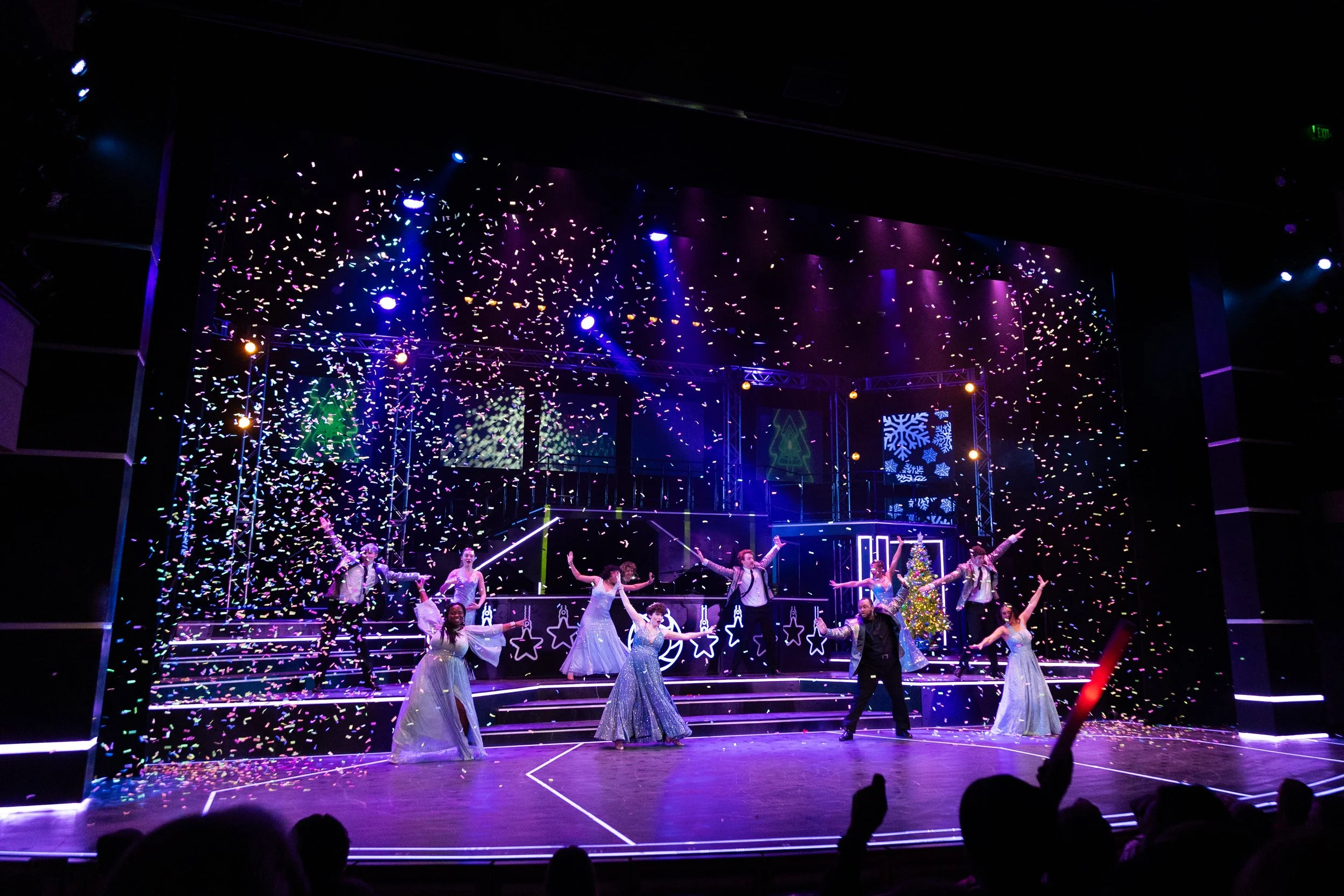 A stage decorated for Christmas with a dance performance, confetti falling, and bright colorful lights. Performers are dressed in festive costumes, with a decorated Christmas tree on the right side.