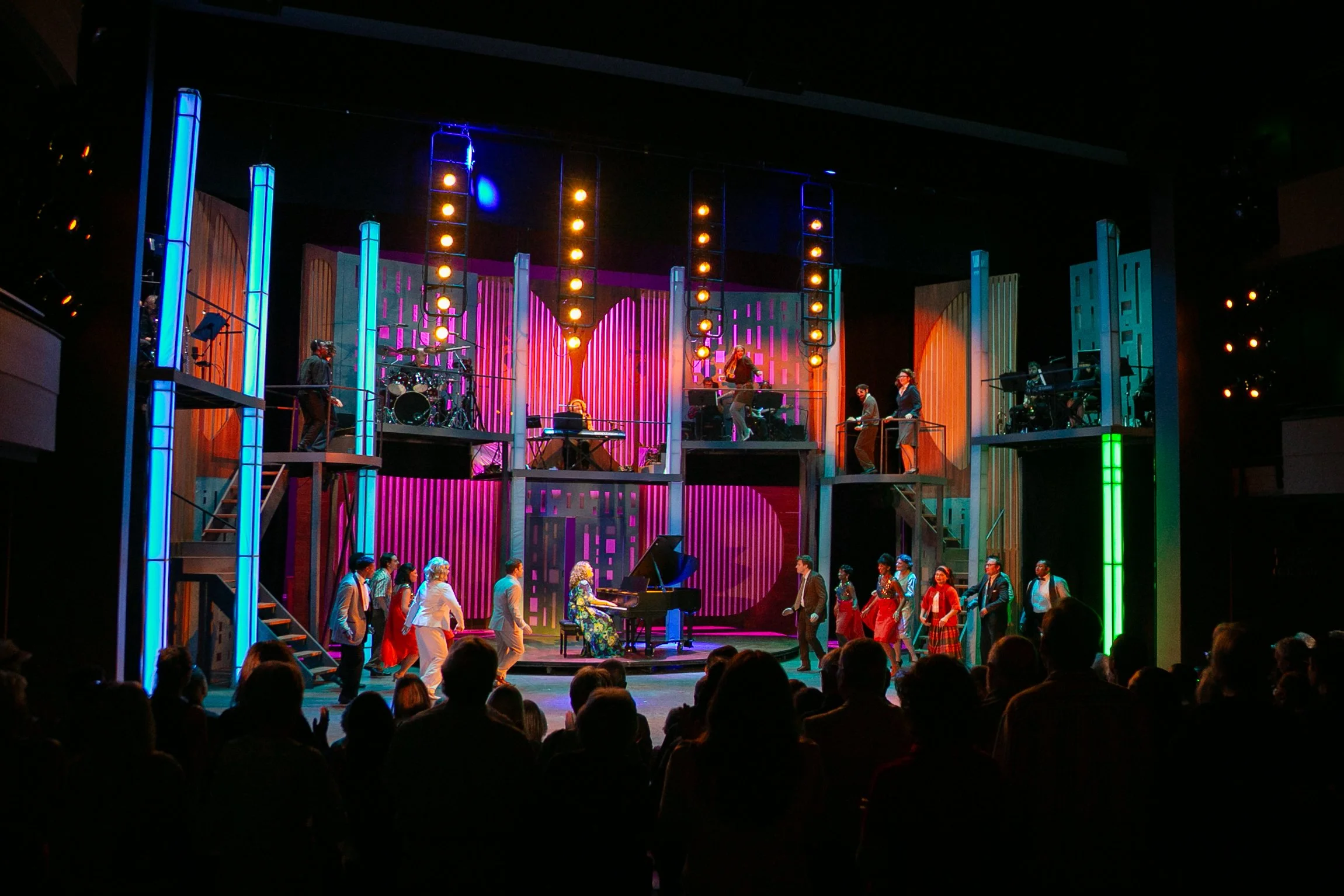 Stage performance with musicians and performers, colorful lighting, and an audience in a theater.