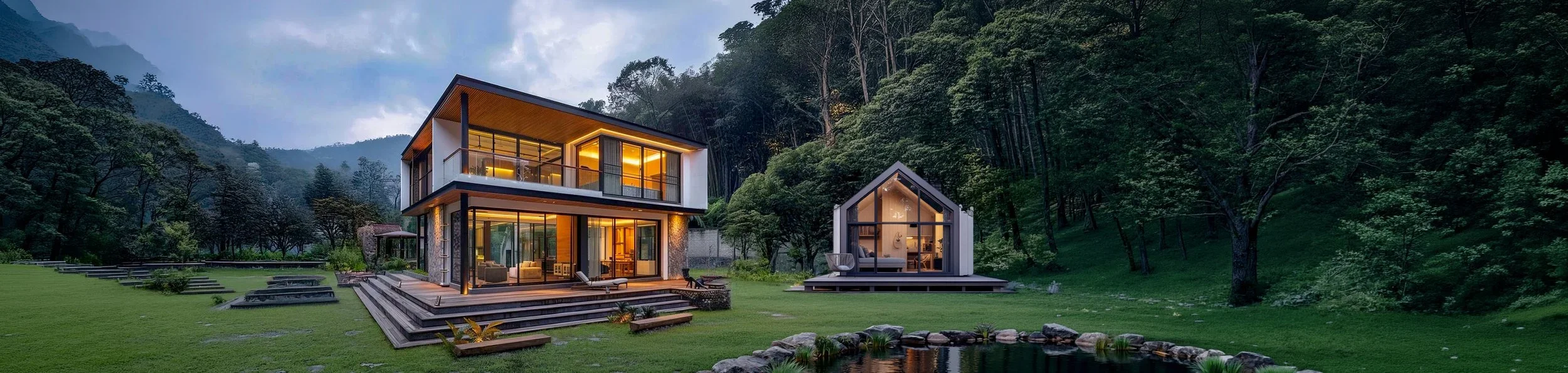 Modern two-story house and a small modern cabin on green lawn with trees and mountains in the background, illuminated at dusk.