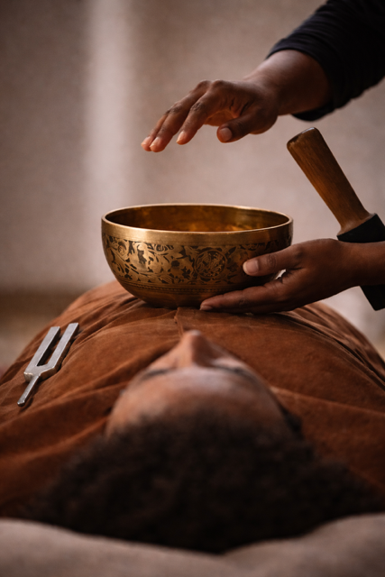 Person lying down with a singing bowl and mallet, receiving sound therapy or meditation treatment.