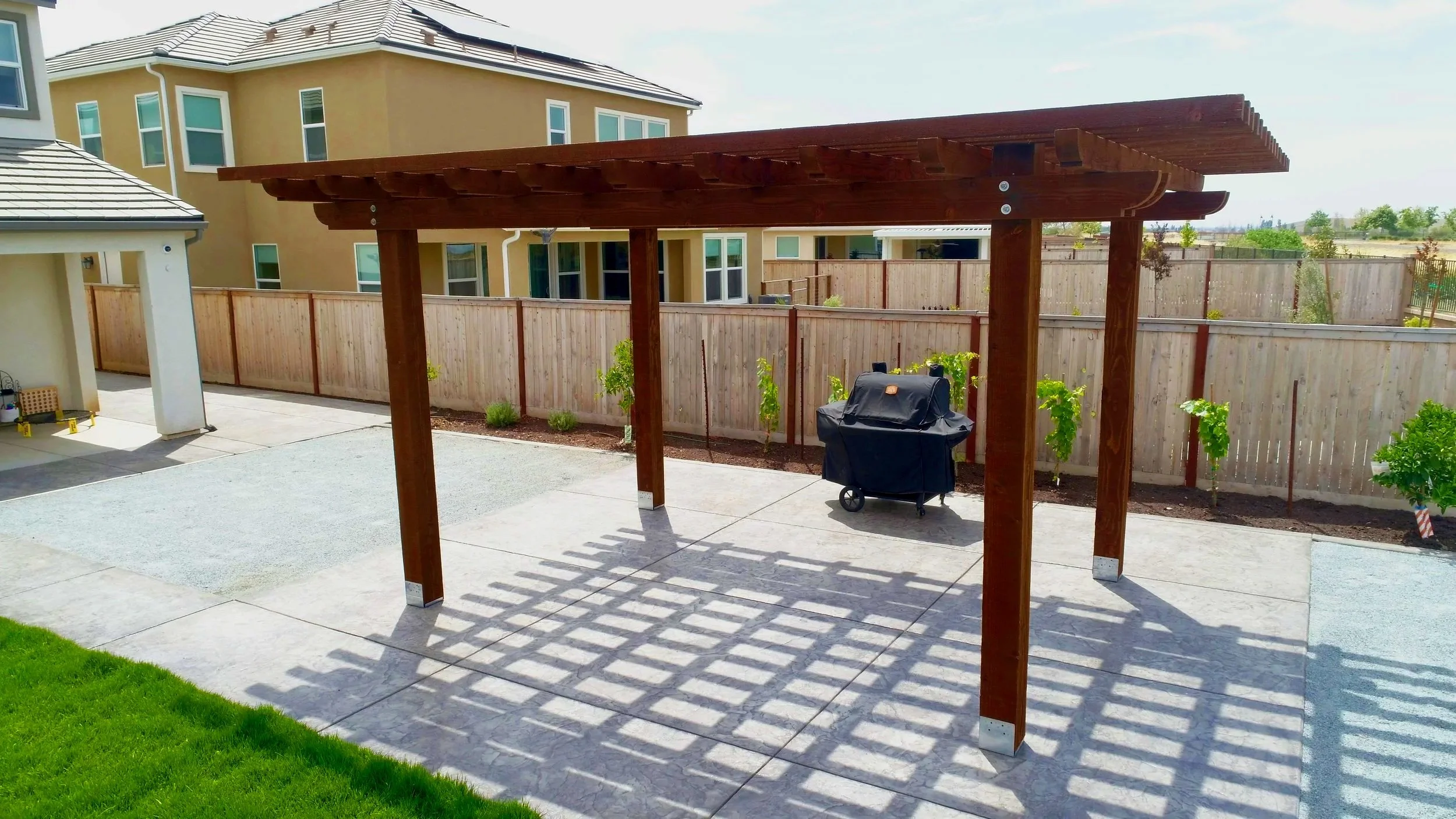 Backyard with a wooden pergola, a barbecue grill, a surrounding wooden fence, small plants, and a house in the background.