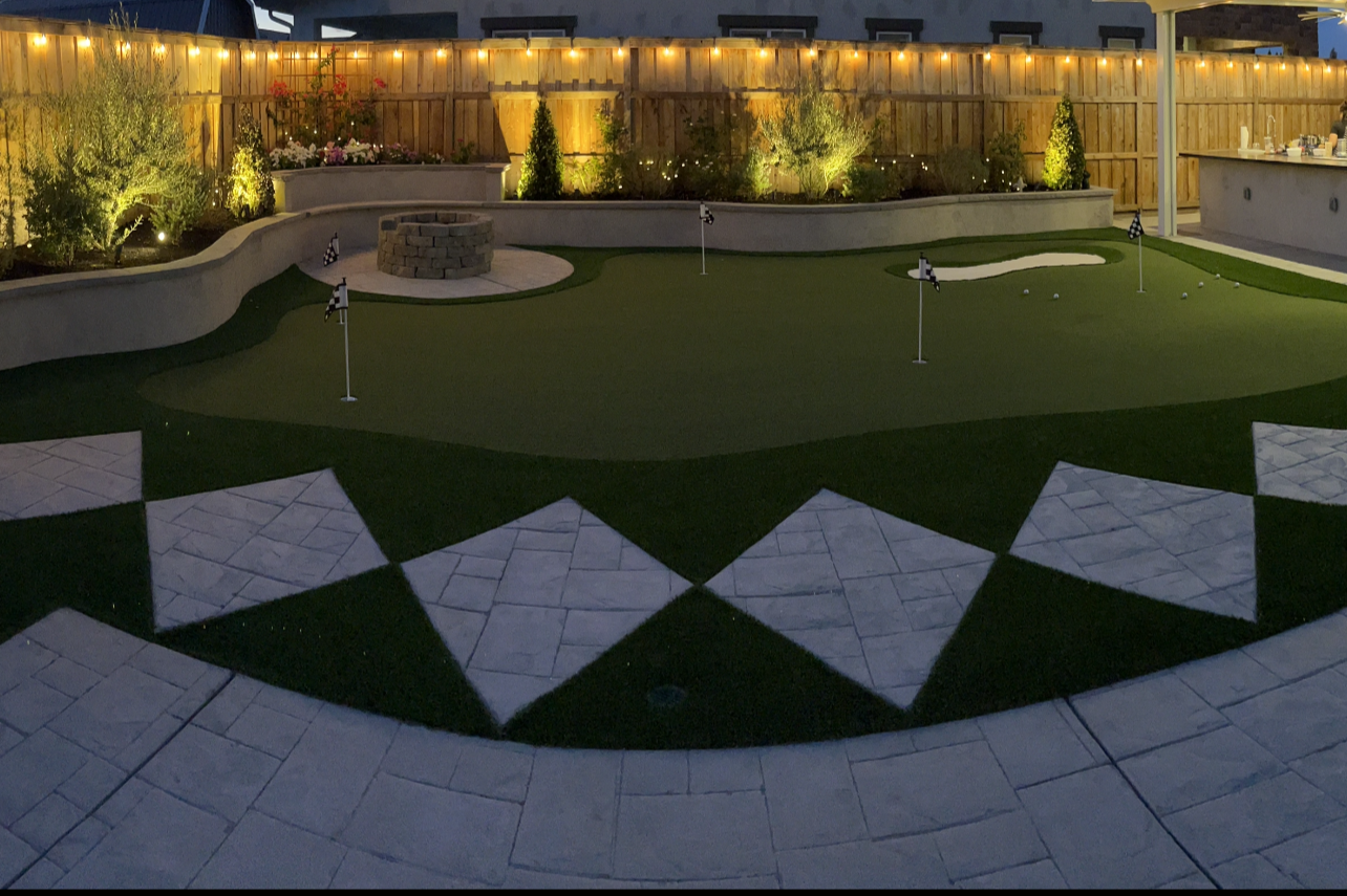 Nighttime backyard with a mini golf course, putting green, sand trap, and flags, illuminated by outdoor lights and surrounded by a wooden fence and landscaped plants.