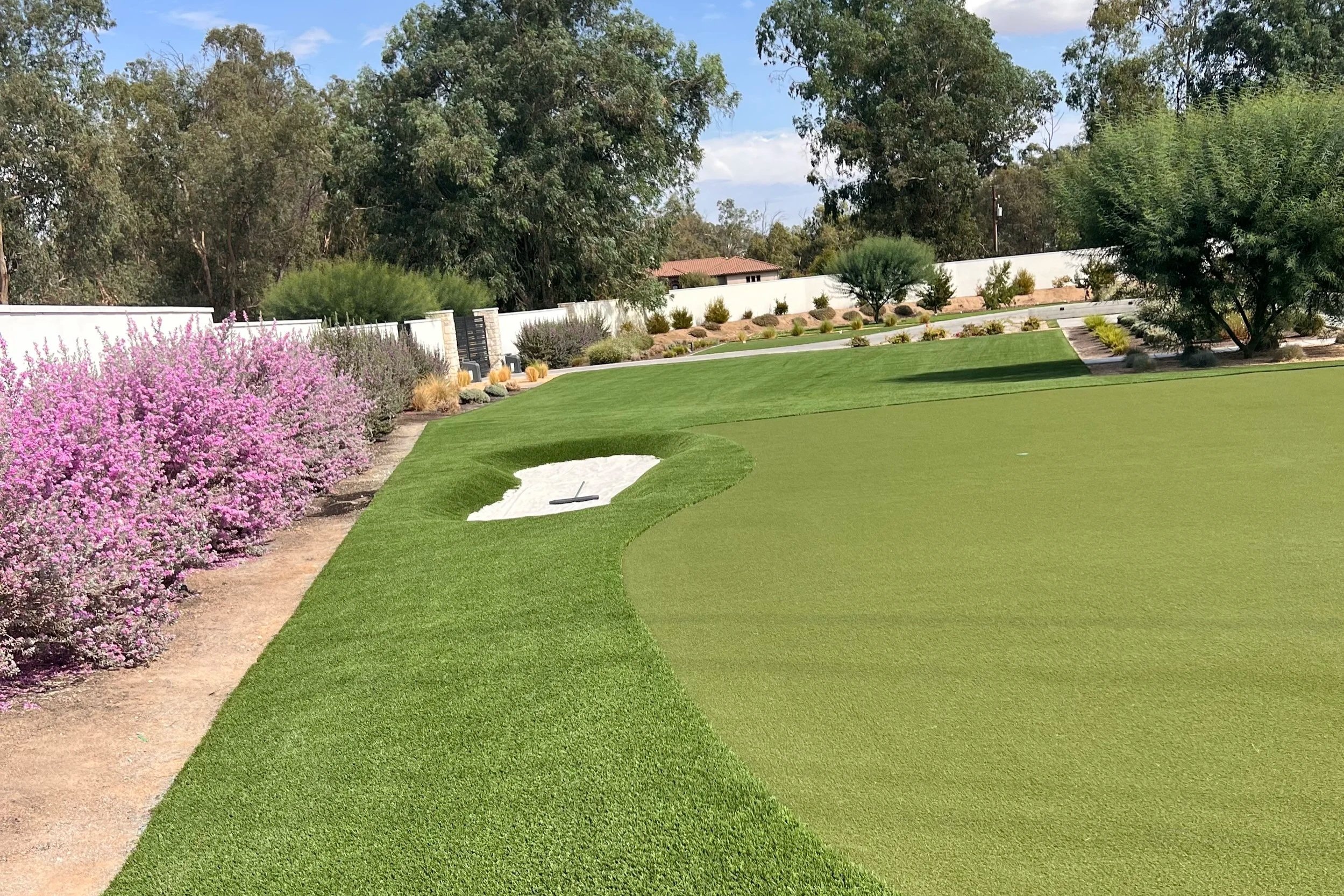 A well-maintained golf course putting green with a sand bunker, surrounded by vibrant pink and purple flowering bushes and trees, under a partly cloudy sky.