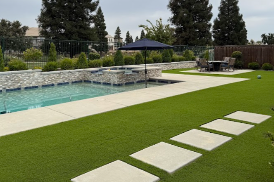 Backyard with a swimming pool, artificial grass, stepping stones, outdoor seating area, and large umbrella.