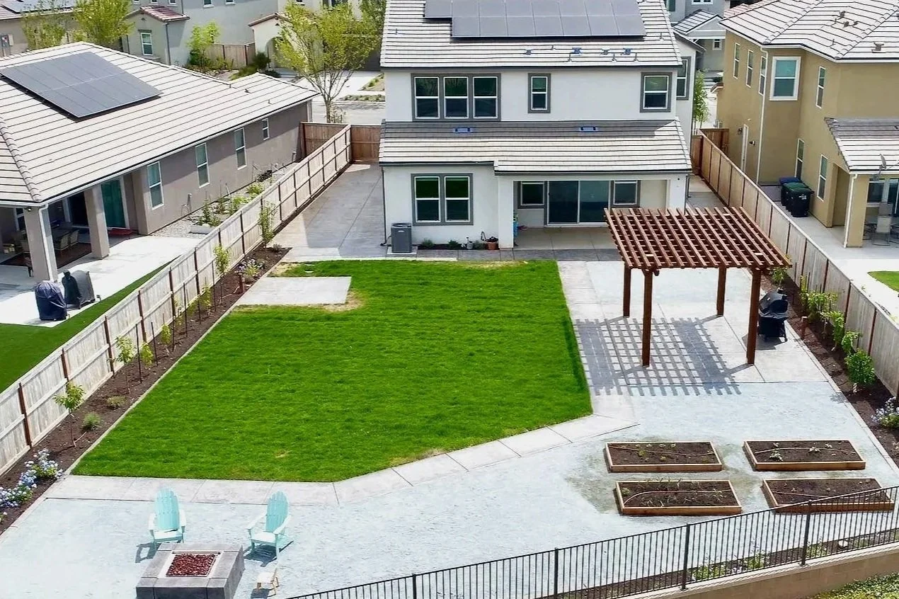 View of a backyard with a central green lawn, a wooden pergola, garden beds, patio area with chairs and a small fire pit, surrounded by a wooden fence and neighboring houses with solar panels on rooftops.