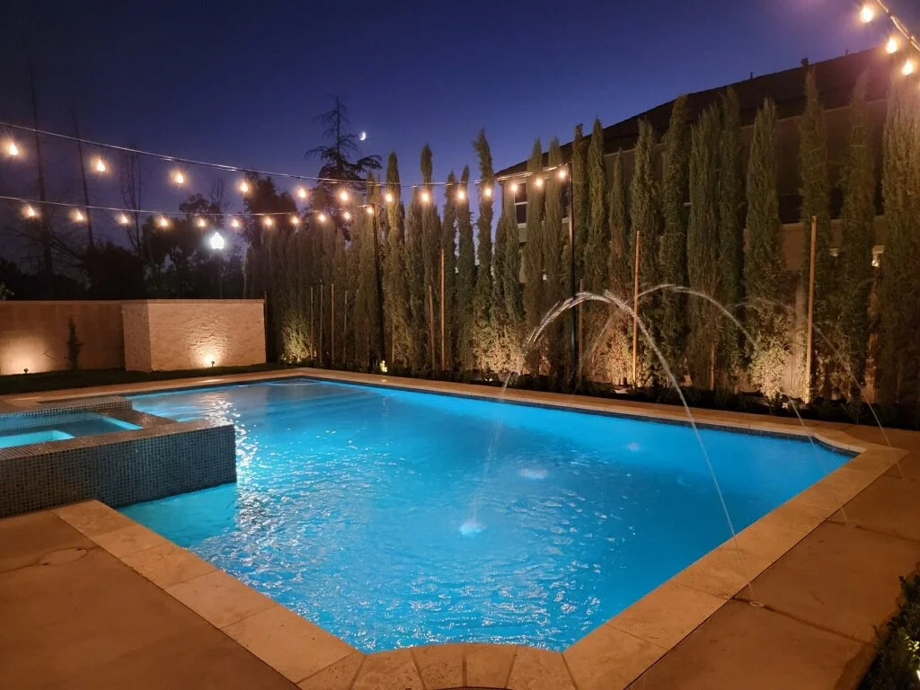 A lit backyard swimming pool with two water fountains at night, surrounded by tall trees, string lights, and a privacy wall.