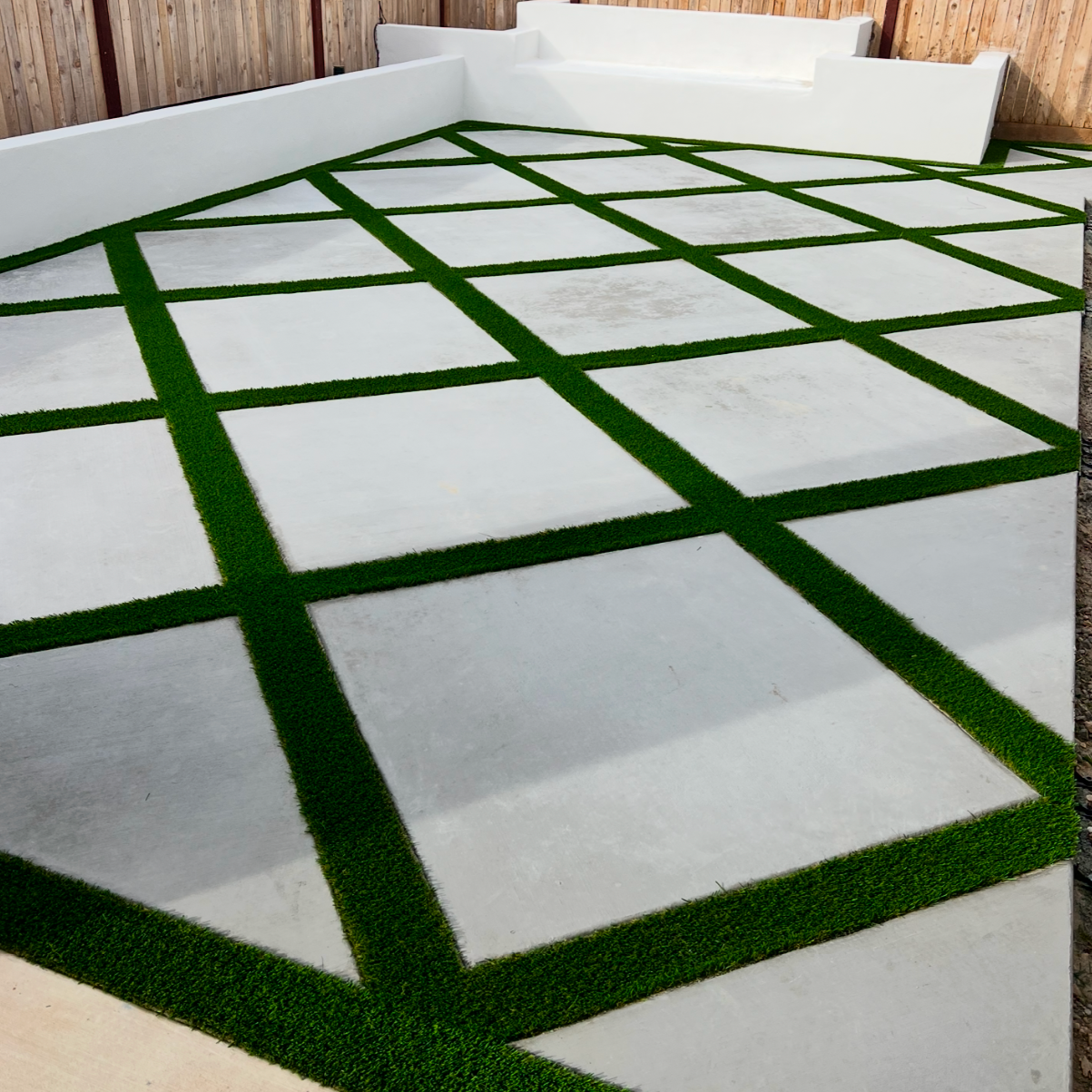 Concrete patio with green artificial grass lines creating a grid pattern, located next to a wooden fence, with a white built-in bench in the corner.