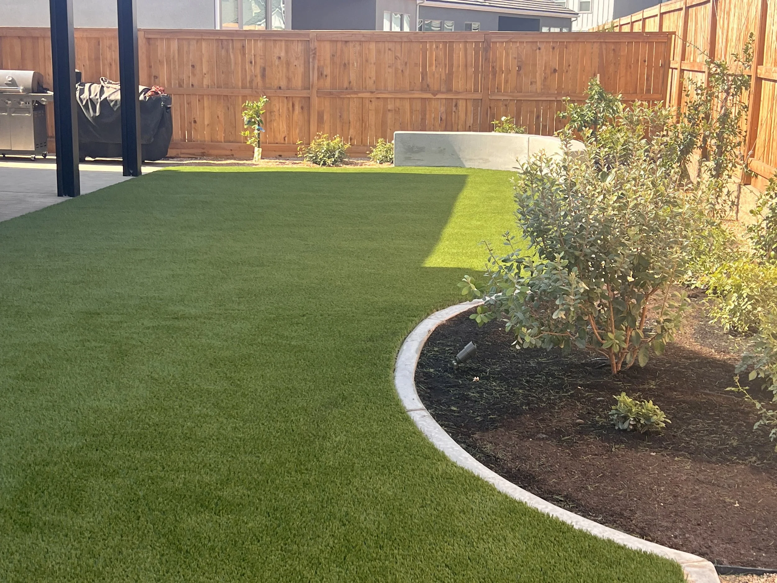 A backyard with a lawn, shrubbery, a wooden fence, and part of a patio with a gas grill.