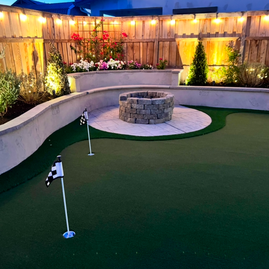 Nighttime backyard with a mini golf putting green, two black-and-white flags, a circular fire pit made of bricks, wooden fencing, string lights, and decorative plants including flowers and small trees.