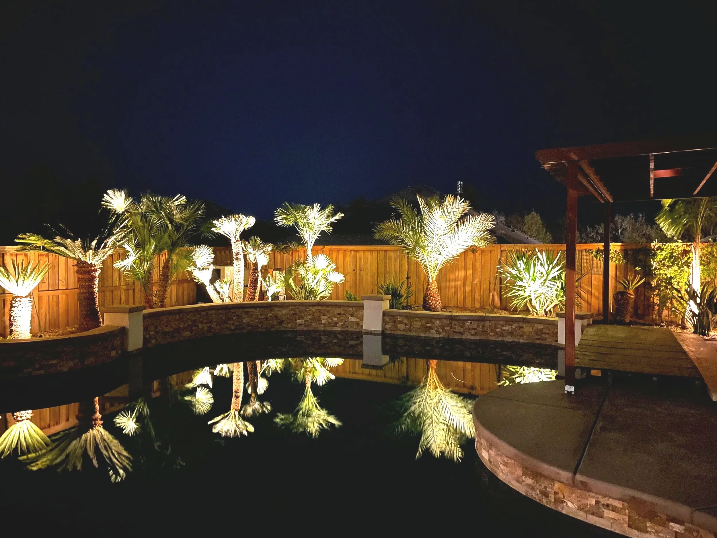 Night view of a backyard with illuminated palm trees, a pool, a wooden fence, and a covered patio area.
