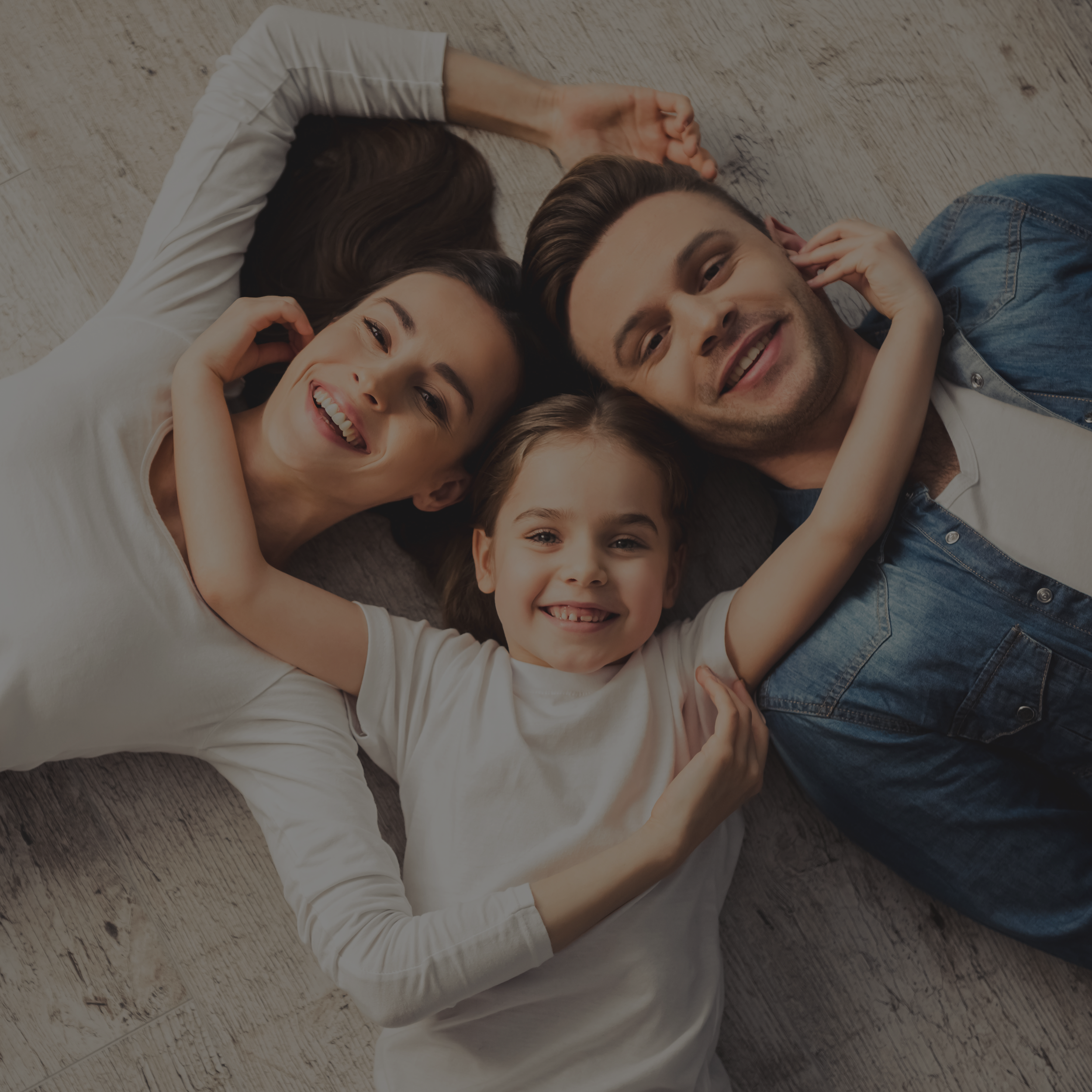Family of four, two adults and two children, lying on wooden floor, smiling and embracing each other.