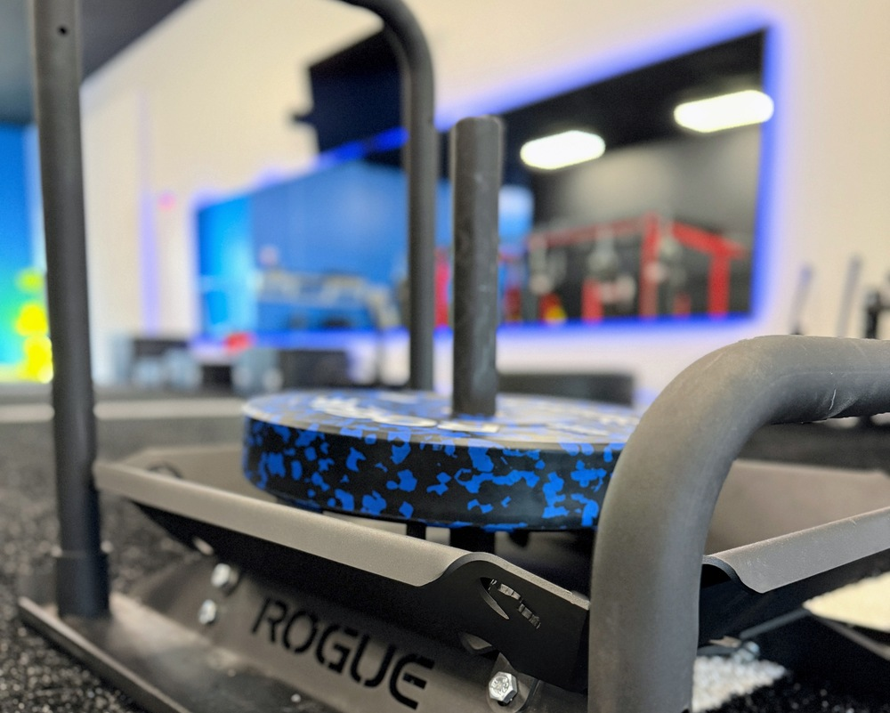 Close-up of a Rogue-branded sled on a black rubber gym floor, with blue and black padded roller in the background at the gym.