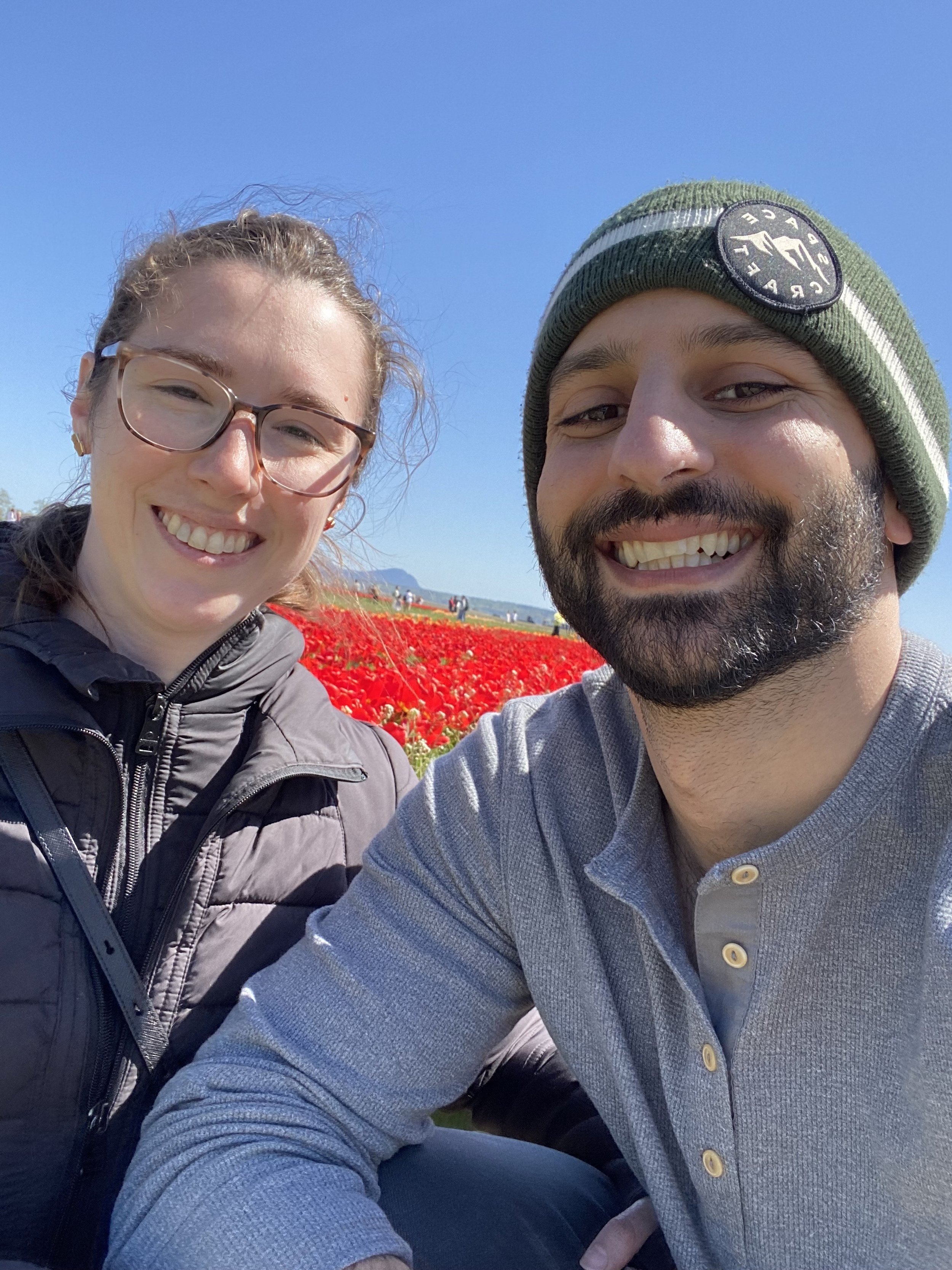 A smiling man and woman taking a selfie outdoors with a field of red flowers behind them under a clear blue sky.