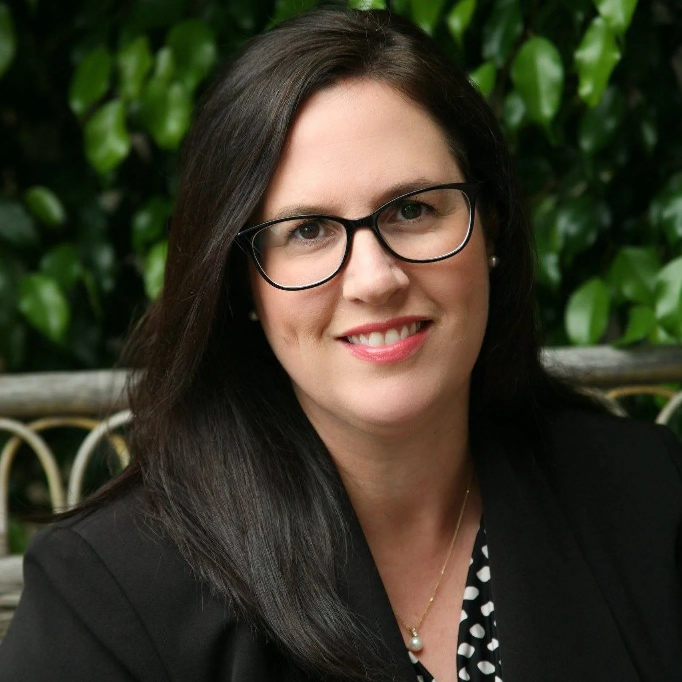 A woman with straight dark hair, wearing glasses, a black blazer, and a pearl necklace, smiling in front of green foliage.