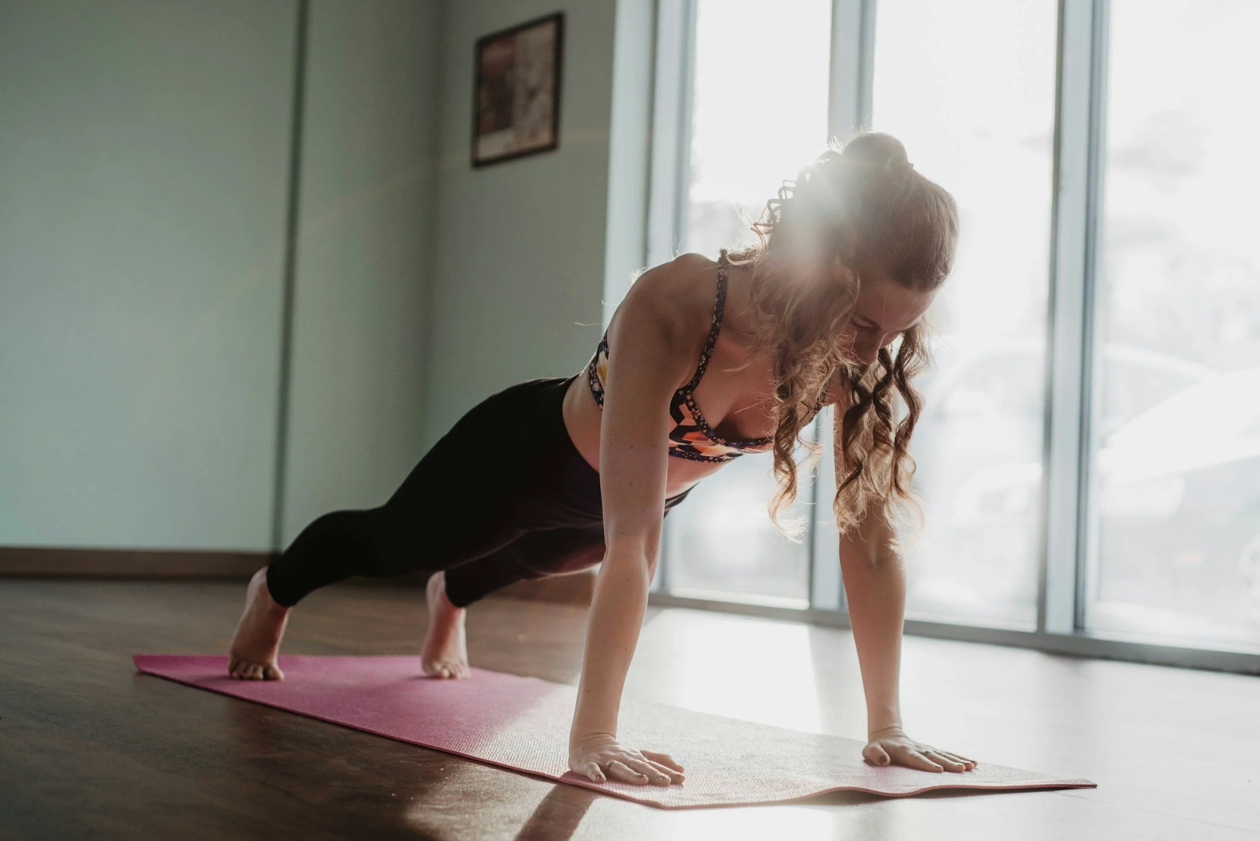 A woman practicing yoga indoors on a pink mat, in a plank position with sunlight streaming through large windows behind her.