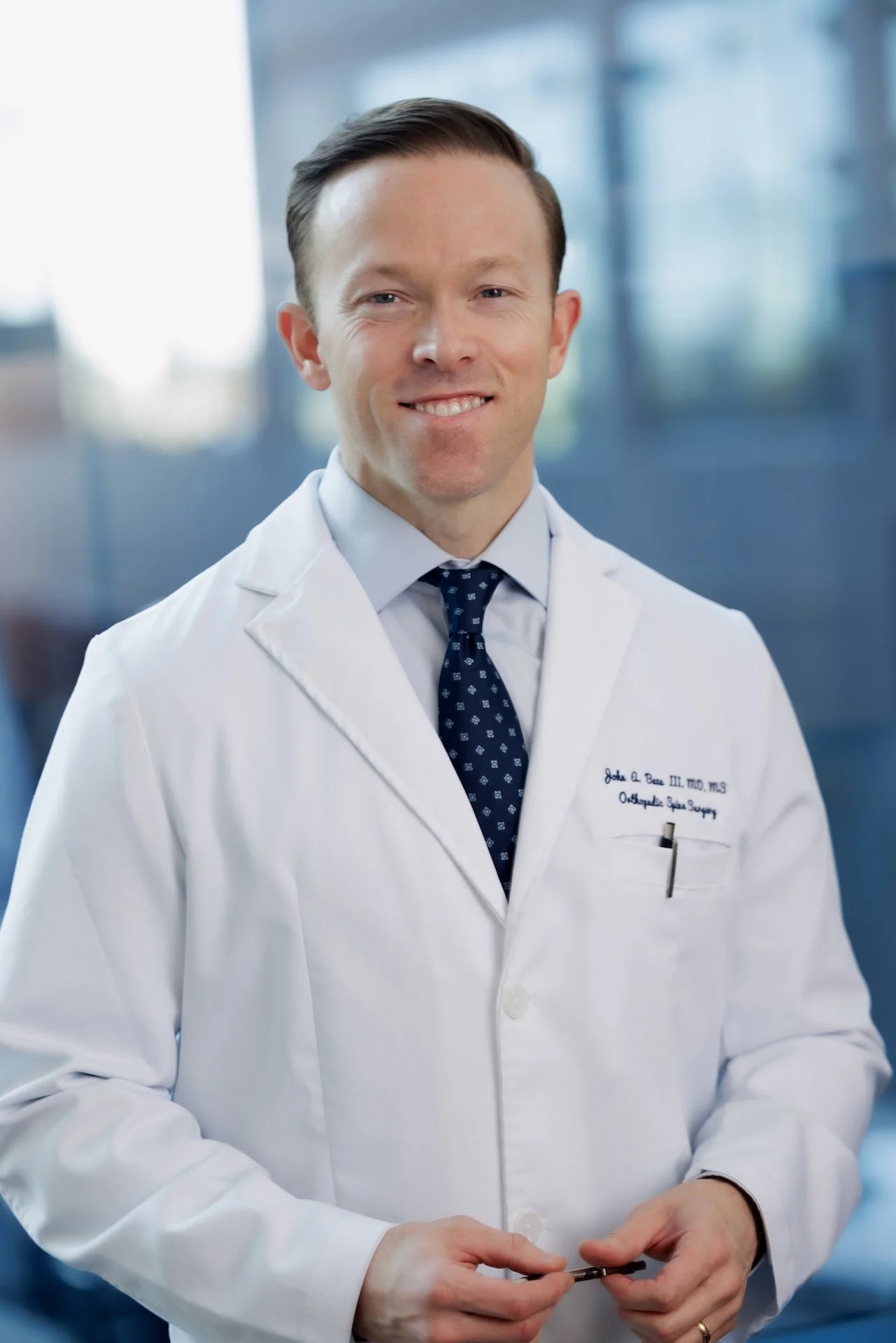 A smiling male doctor in a white lab coat standing in a modern medical facility.