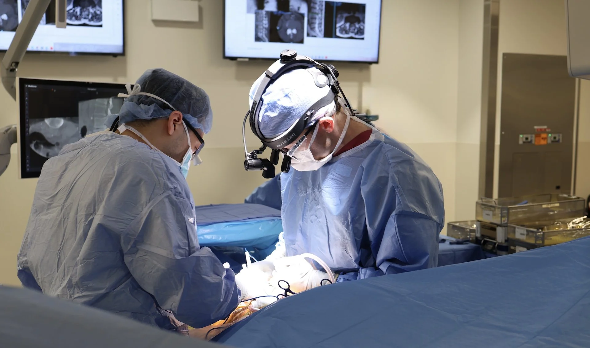 Two surgeons performing an operation in an operating room, wearing scrubs, masks, and surgical gloves, with medical monitors displaying medical images in the background.