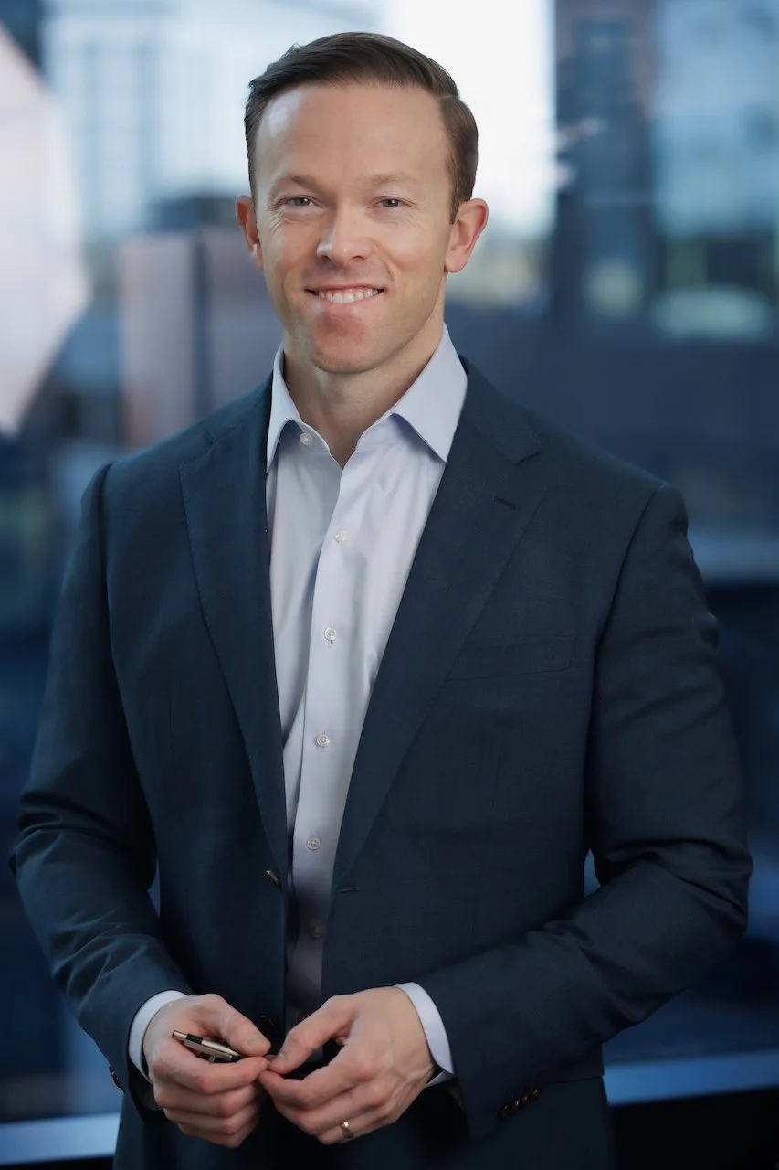 A professional man in a dark suit and white shirt smiling and holding a pen, standing in an office with large windows.