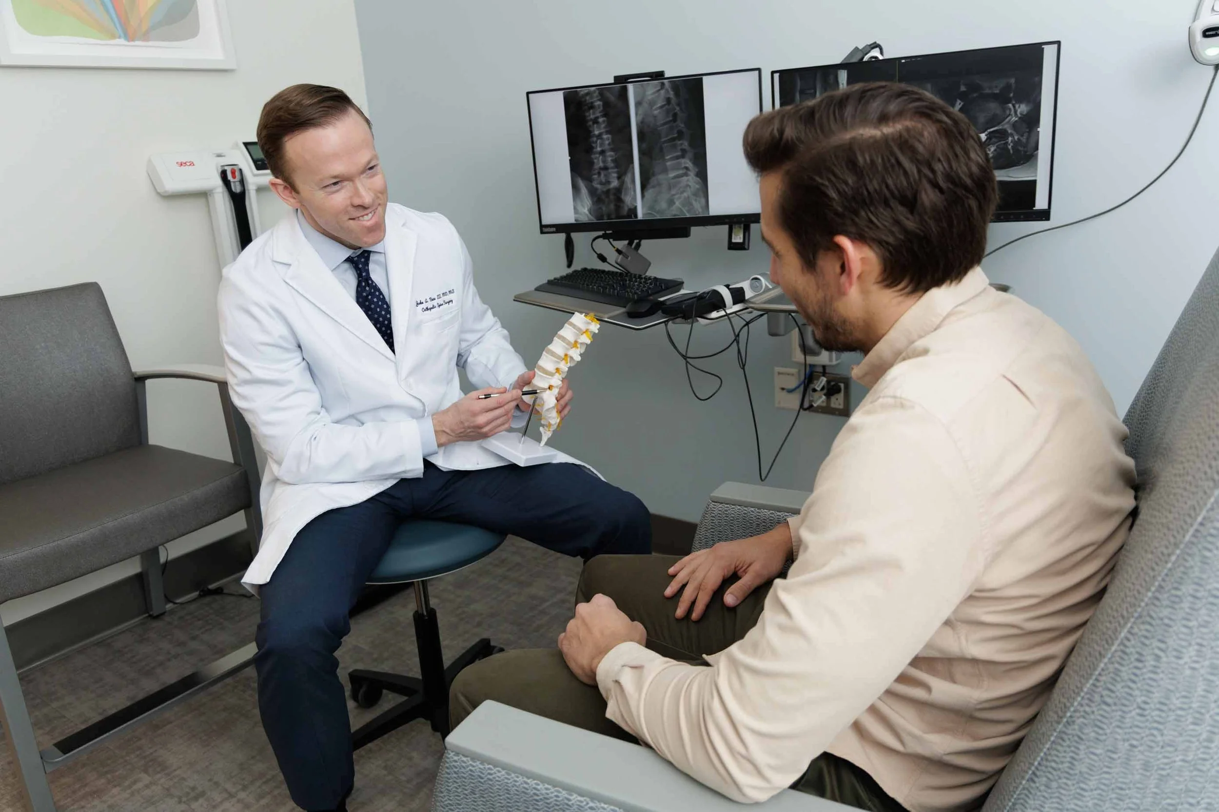Doctor showing a spinal model to a patient in a medical office with medical imaging screens in the background.