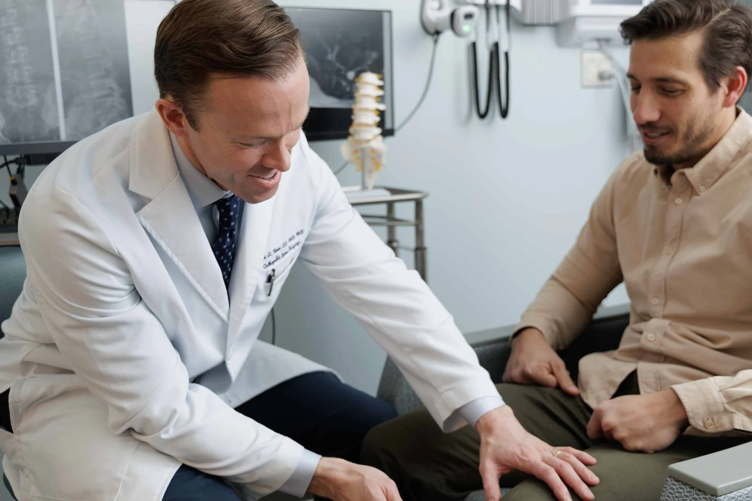 A doctor smiling and examining a patient's leg in a medical office, with medical equipment and anatomical models in the background.