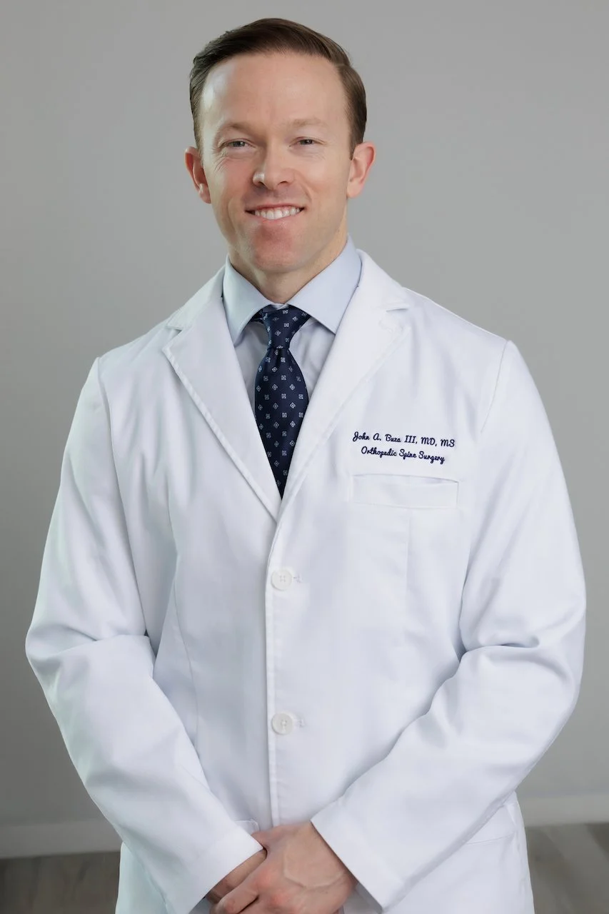Professional portrait of a male doctor in a white lab coat with embroidered name and specialty, wearing a light blue dress shirt and a navy blue patterned tie, standing against a plain gray background.
