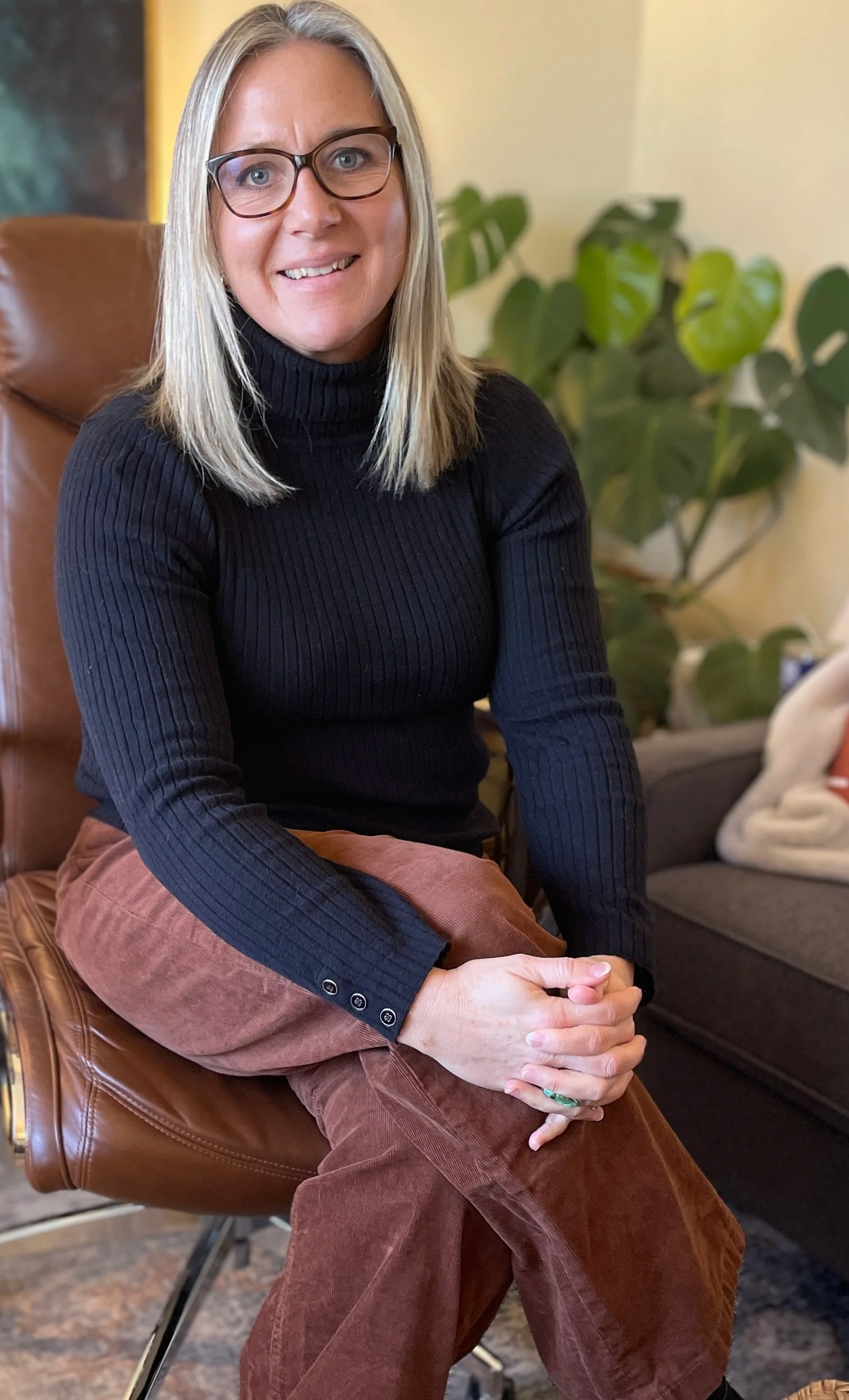 Karin Lyon, wearing glasses, a black turtleneck sweater and brown corduroy pants, sitting in a mid-century modern chair with her hands clasped in her lap. In the background, there is a large green houseplant and a gray sofa.