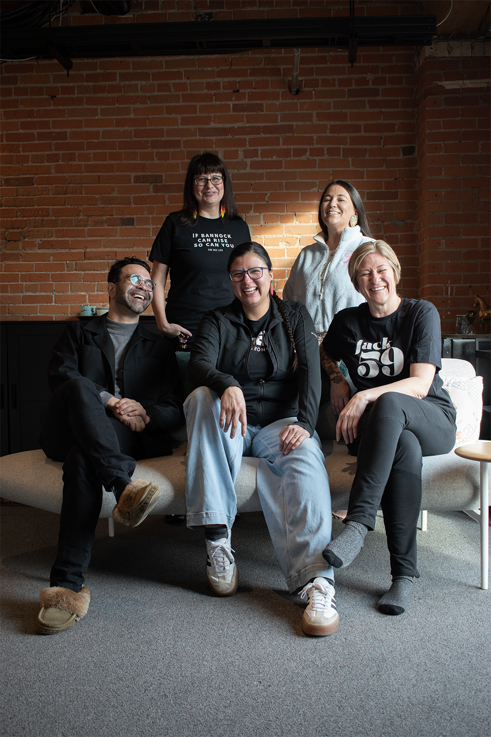 Group of five diverse people sitting and standing on a beige couch in a room with a brick wall background, smiling and laughing together.