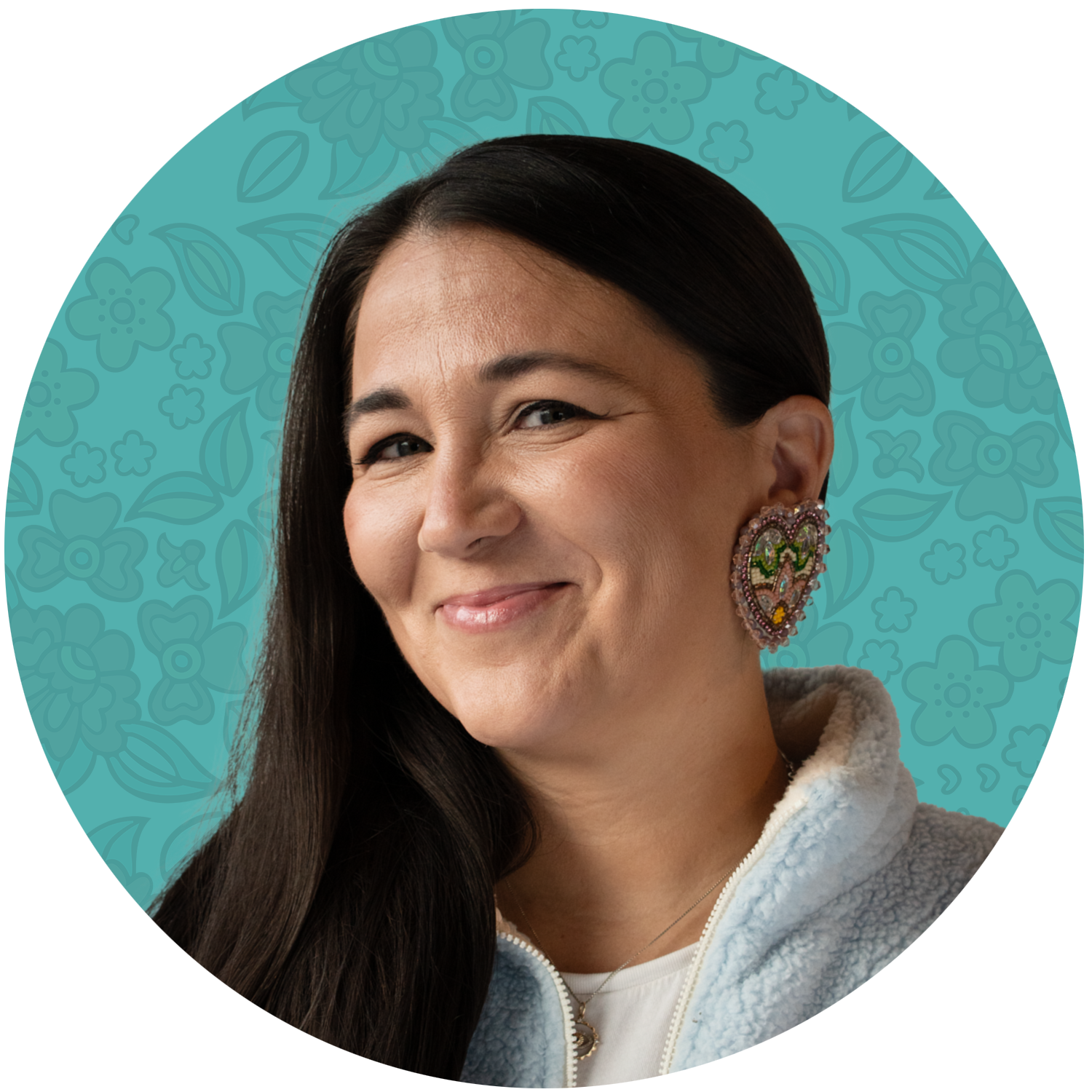 Shani Gwin. Founder, pipikwan pêhtâkwan & wâsikan kisewâtisiwin. A woman with long dark hair, smiling, wearing colorful earrings and a white fleece jacket, with a floral patterned blue background.