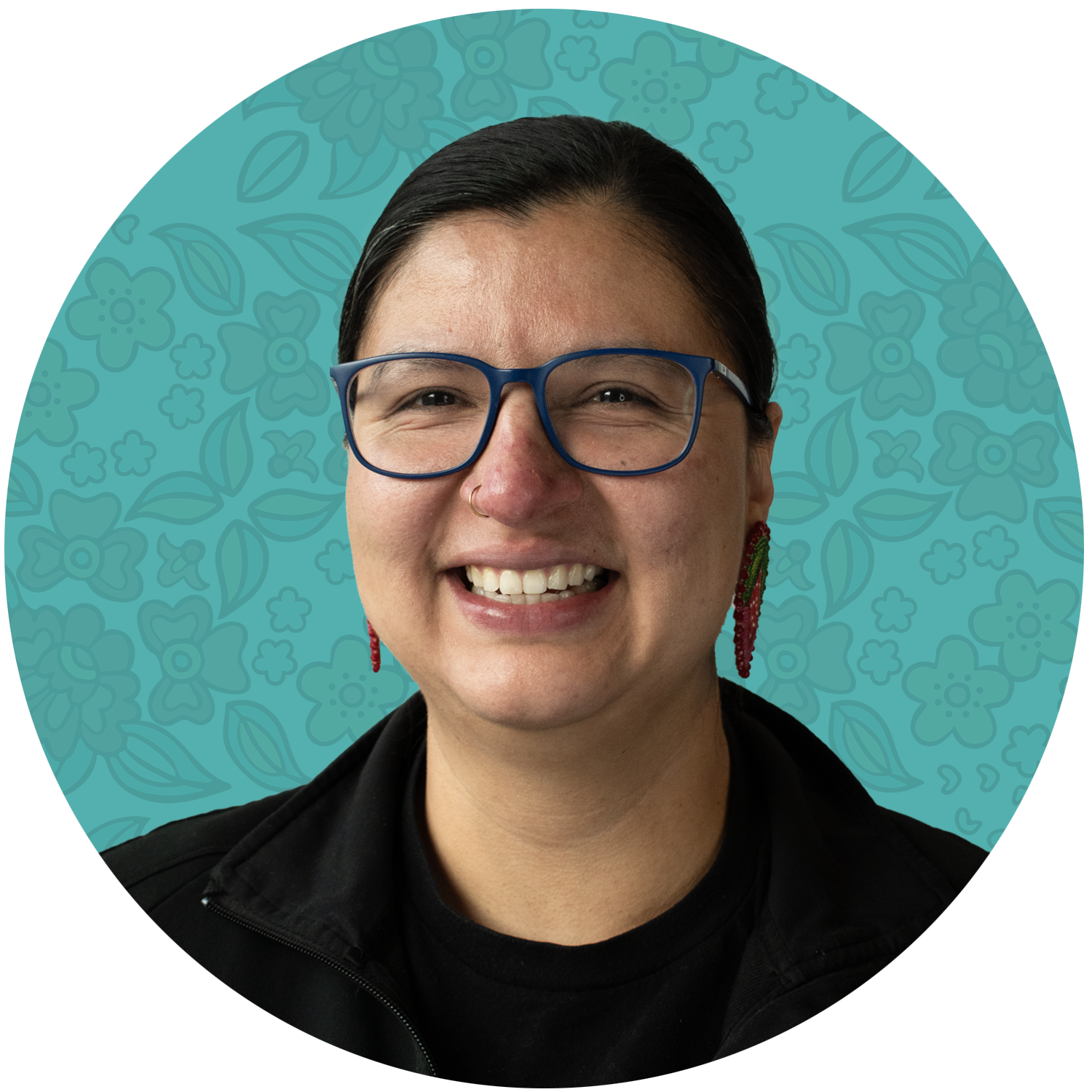 Mallory Yawnghwe. Indigenous Box Foudner. Close-up portrait of a smiling person wearing glasses with a blue frame, a nose ring, and colorful earrings, against a teal background with a floral pattern.
