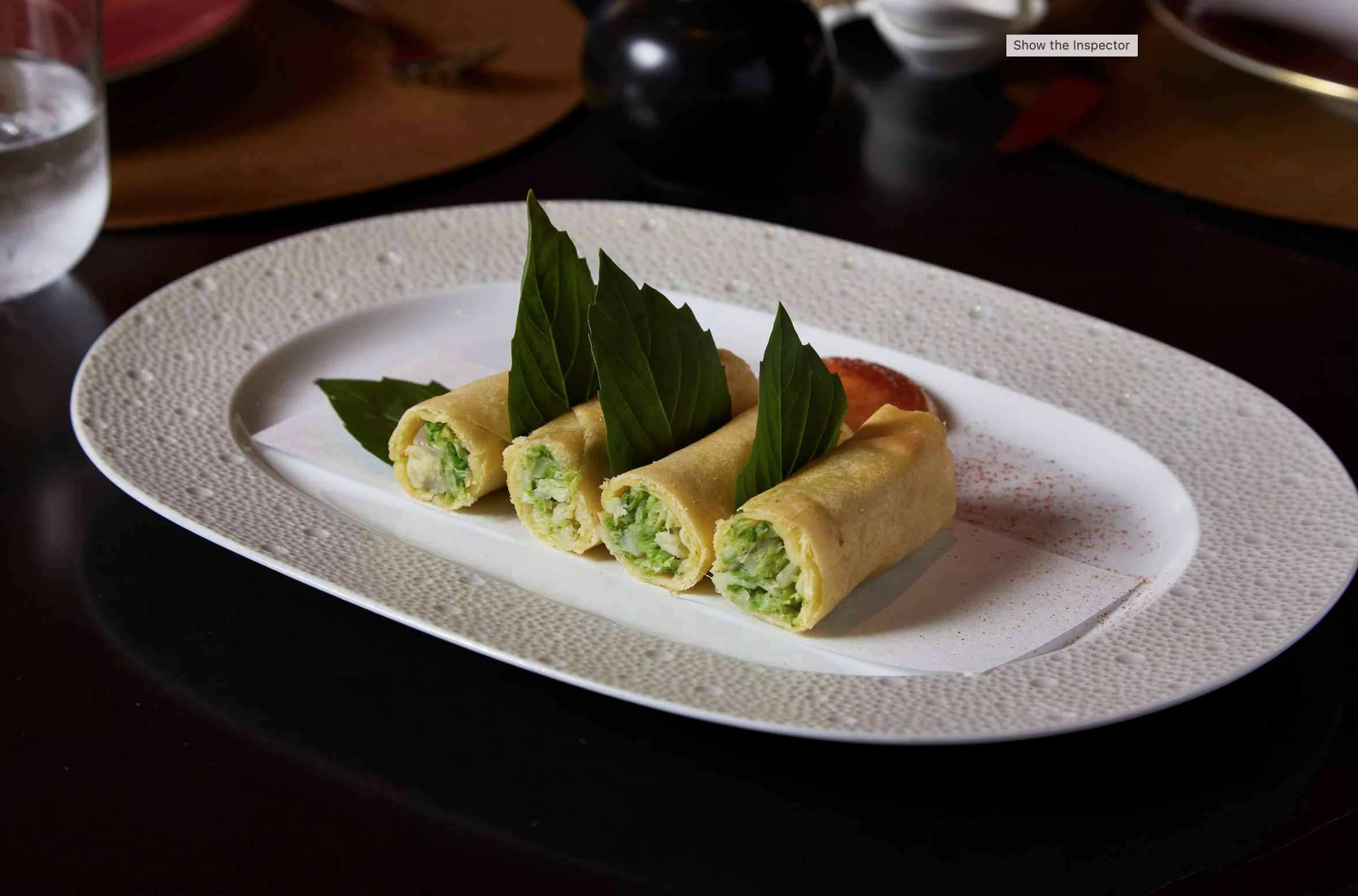 Plate with four spring rolls garnished with fresh green basil leaves, set on a decorative white plate. In the background, a glass of water and some dark-colored dishes are visible. Robert Ranfranz branding, design and ecommerce.