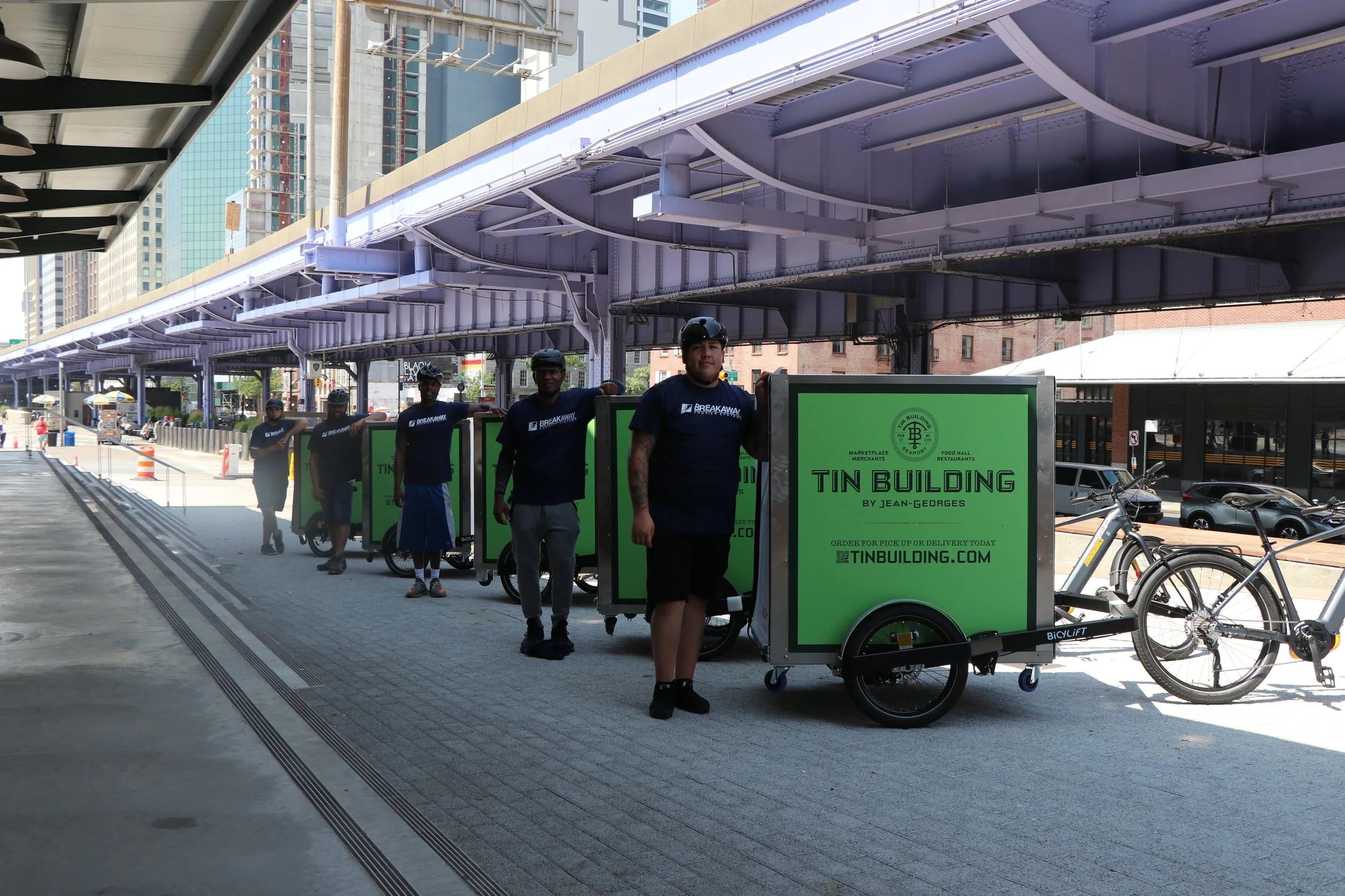Four workers in black helmets and matching shirts standing in line with green and black delivery carts under an elevated train track in an urban area. Robert Ranfranz branding, design and ecommerce.