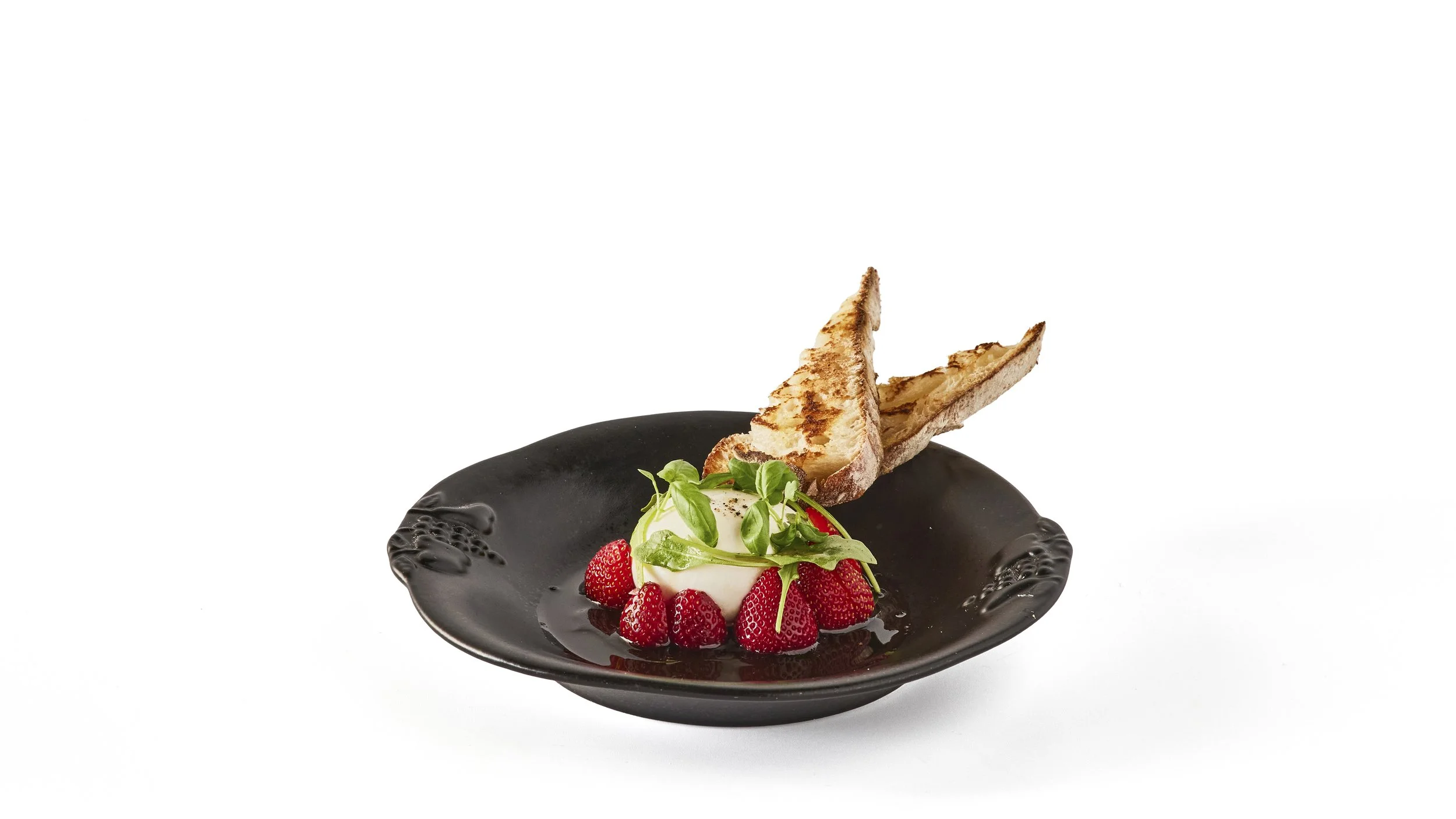 A dessert featuring a black ceramic plate with a dome of white cream surrounded by fresh strawberries, decorated with green microgreens and two slices of toasted bread. Robert Ranfranz branding, design and ecommerce.