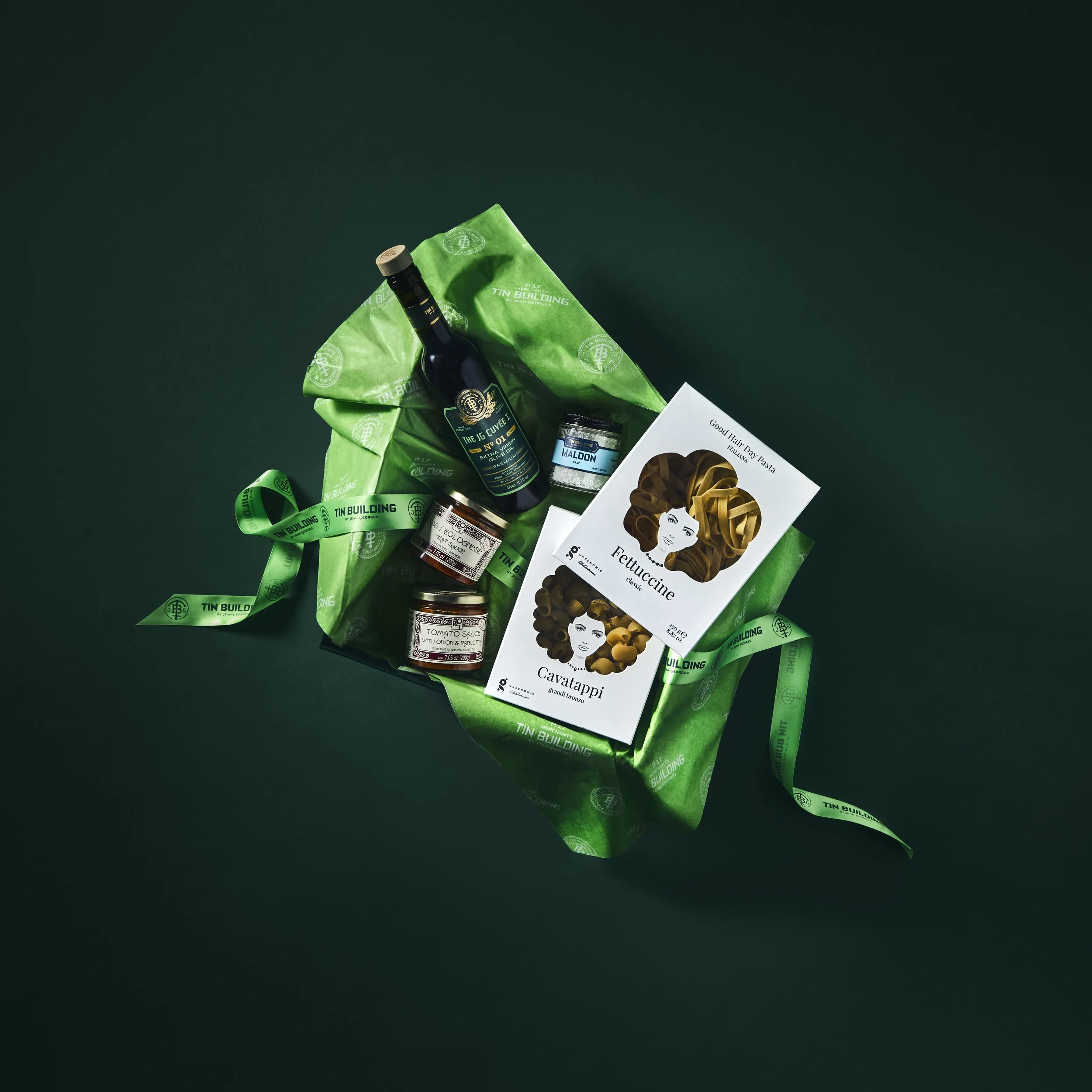 Gift basket with green tissue paper, a bottle of red wine, jars of pasta sauce, a jar of Maldon sea salt, and pasta boxes of Fettuccine and Cavatappi.